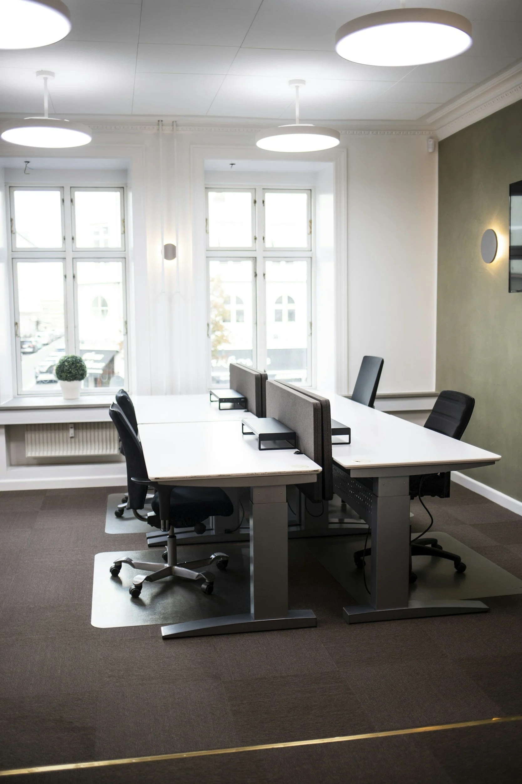 Ein modernes Büro mit weißen Tischen, schwarzen Bürostühlen, Fenstern mit natürlichem Licht, und einer grünen Wand auf der rechten Seite.
