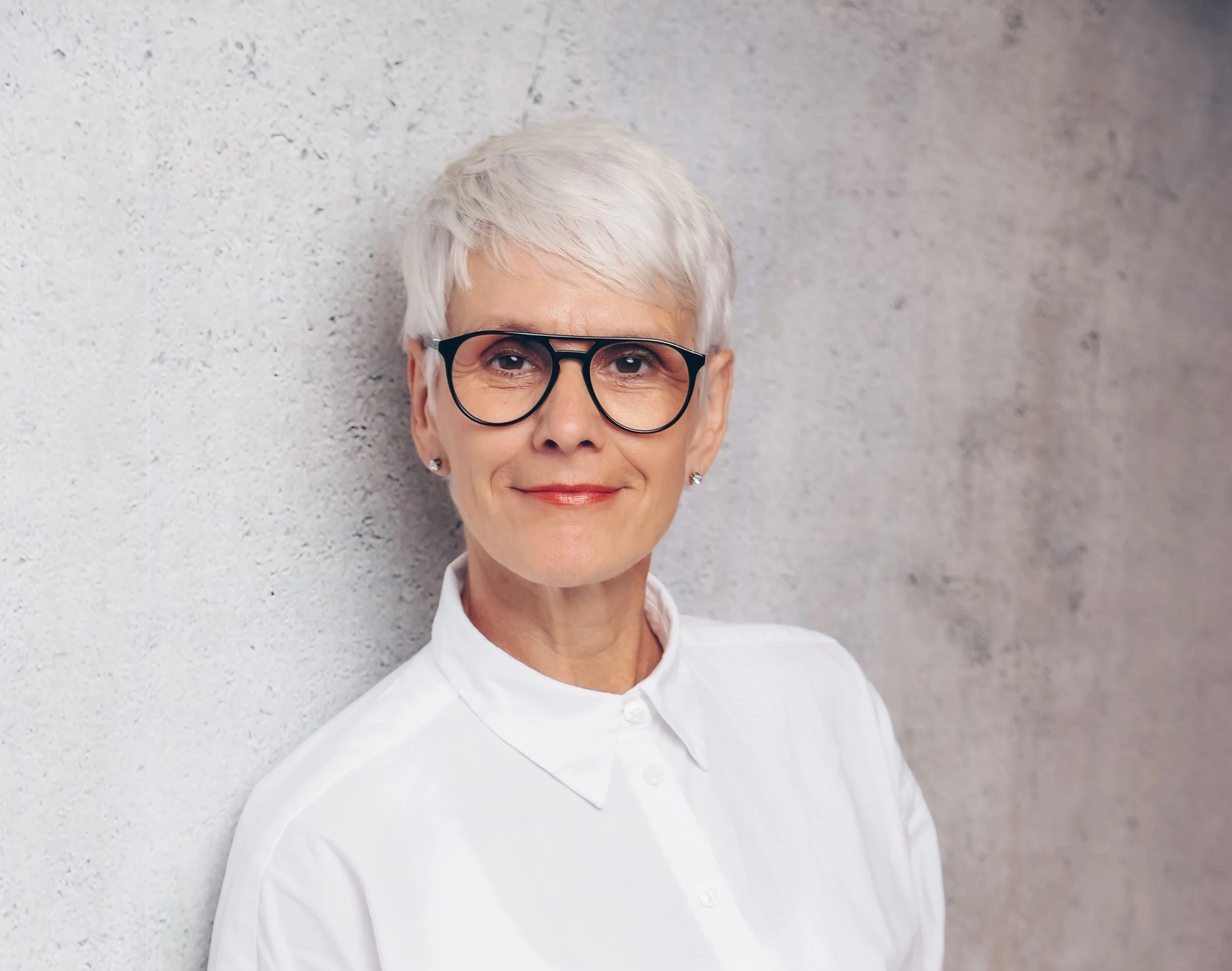 Porträt einer älteren Frau mit kurzen weißen Haaren, Brille und weißen Hemd, vor einer grauen Wand, lächelnd.