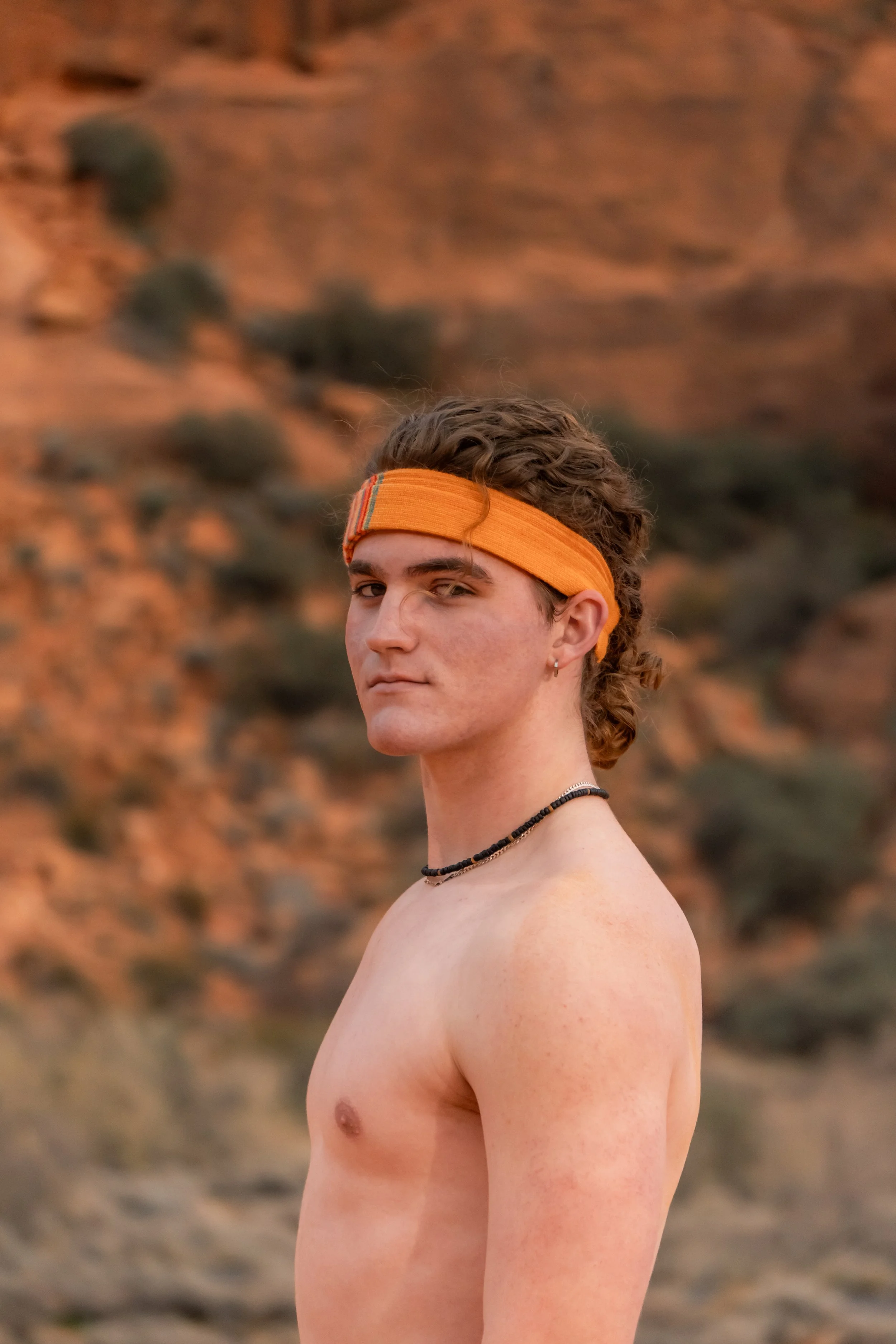 A young man with curly brown hair wearing an orange headband standing shirtless outdoors with a blurred desert landscape in the background.