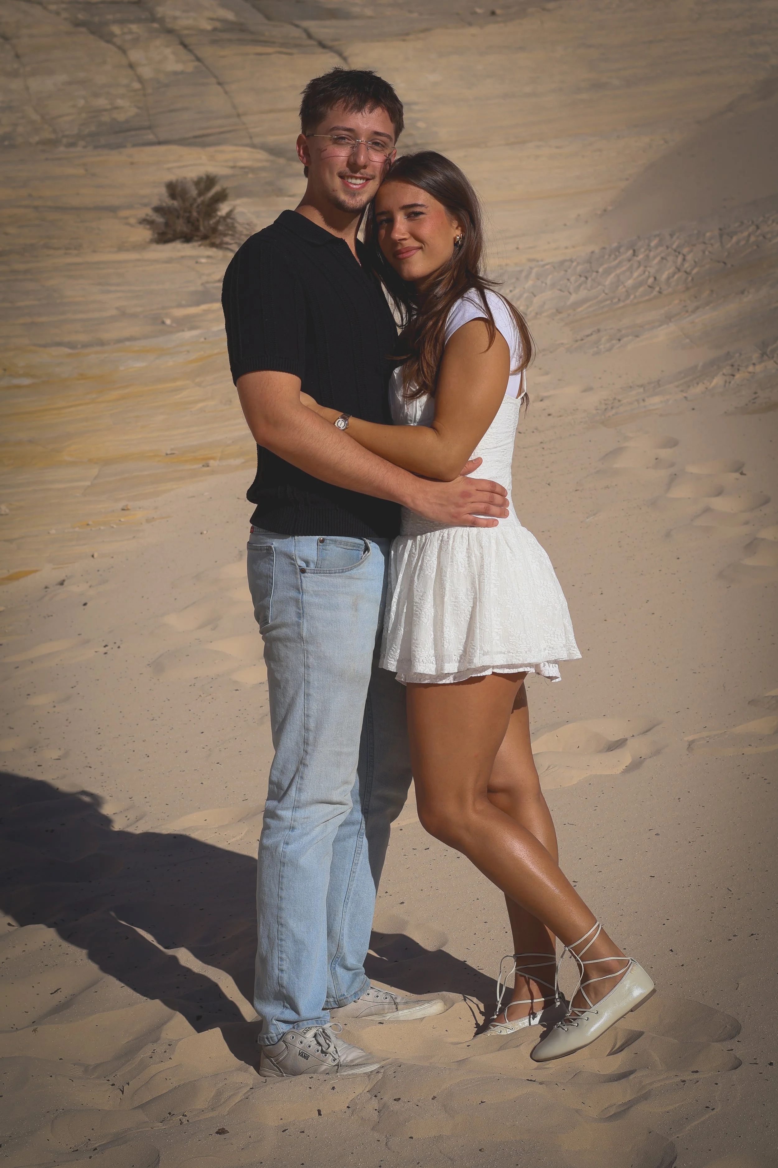 A young couple standing close together on a sandy desert with a rocky, canyon-like background. The man is wearing a black short-sleeve shirt, light blue jeans, and white sneakers, and has glasses. The woman is dressed in a white sleeveless top, a white skirt, and white platform shoes with ankle straps. They are smiling and embracing each other.