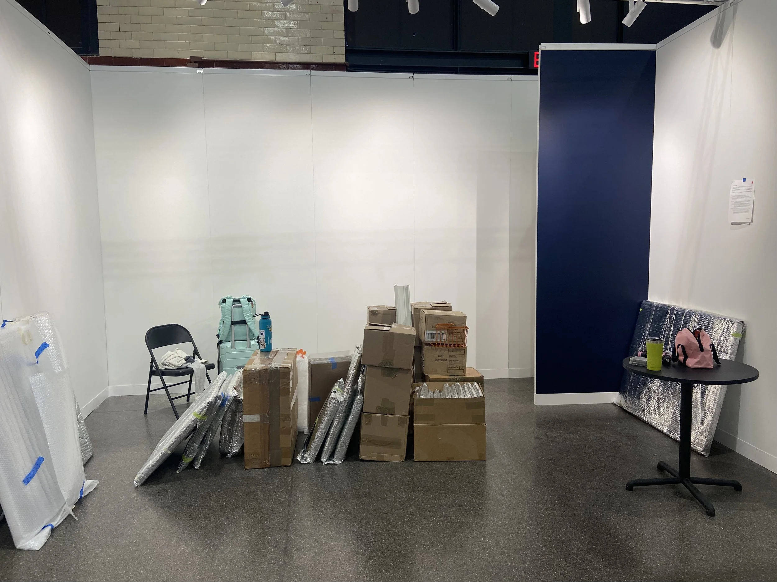 Empty art gallery with walls, scattered cardboard boxes, rolled-up posters or artwork, a chair, a table with bags, and various items for installation or display.