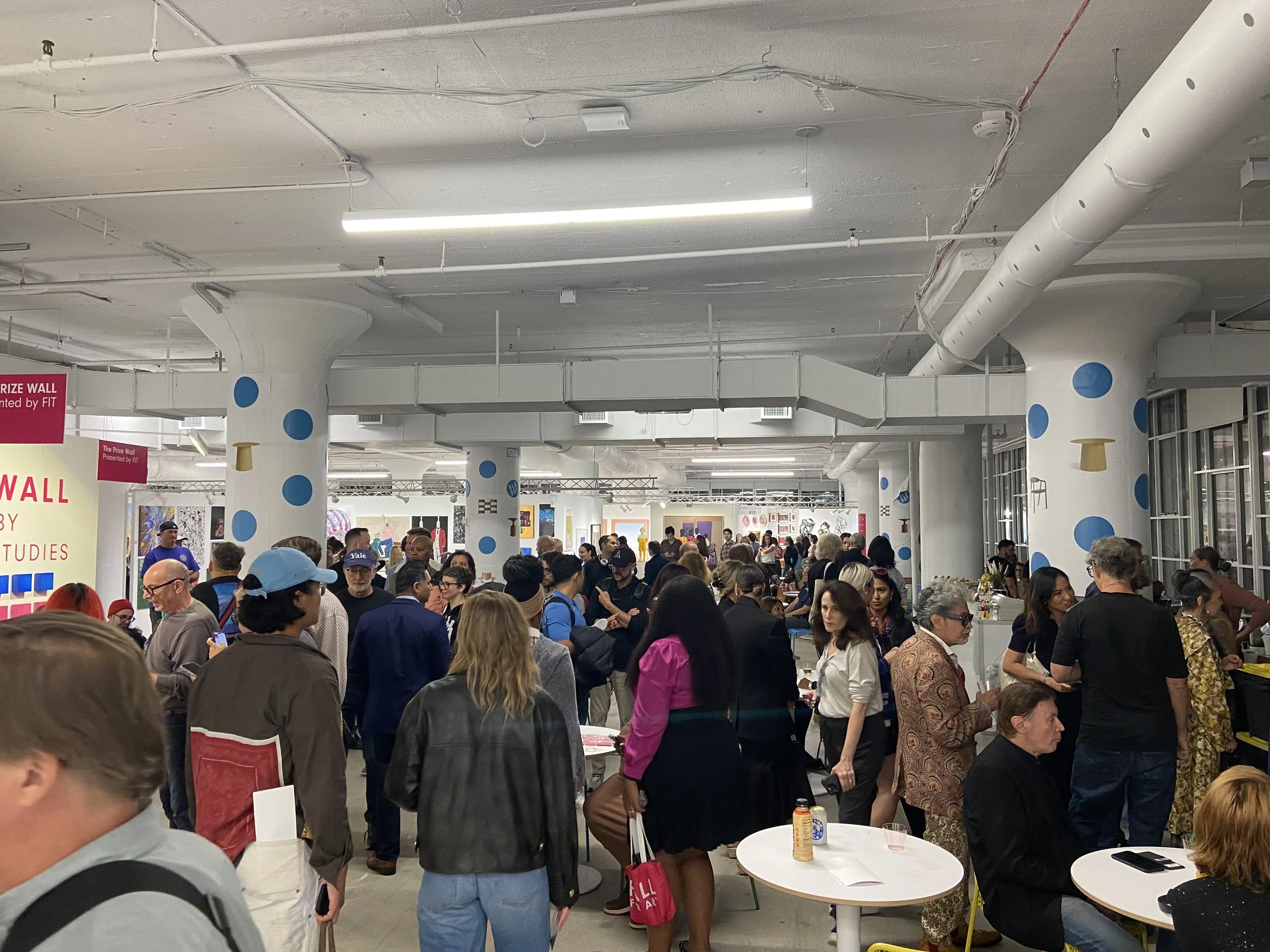 A large indoor art exhibition or auction space filled with a diverse crowd of people walking and browsing various artworks. The ceiling has visible pipes and ducts, with bright lighting.