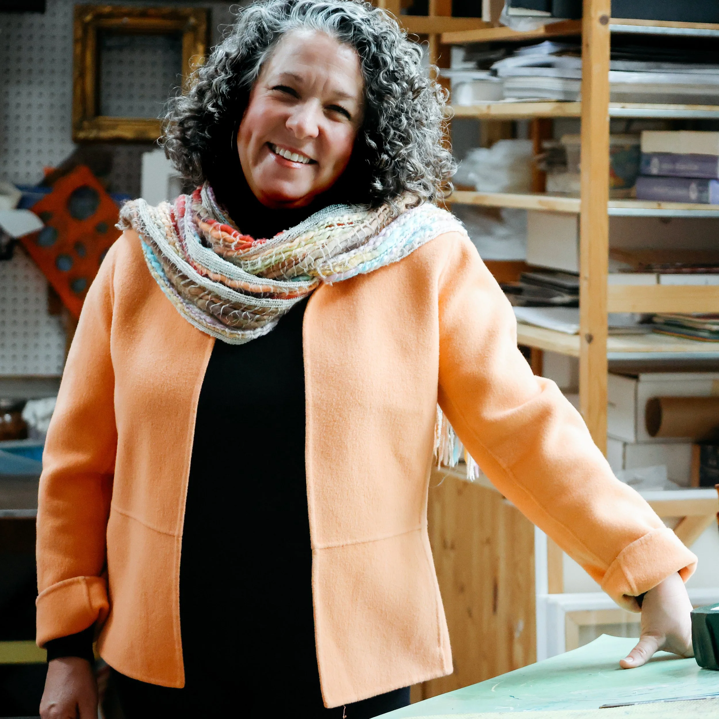 Jill J Galloway, with curly gray hair smiling, wearing an orange coat and a multicolored scarf, standing in a room with shelves filled with papers and books.