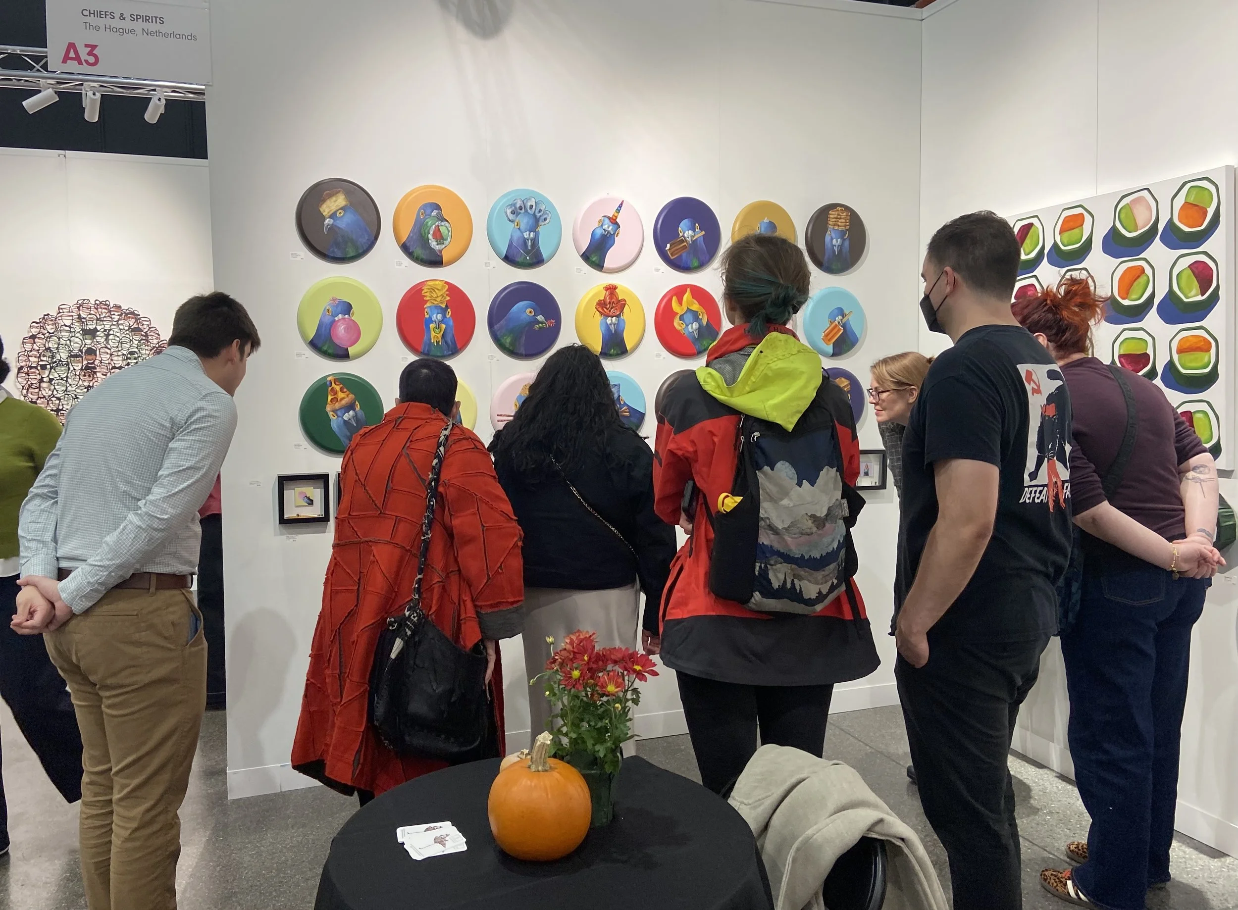 People gathered at an art exhibition with colorful portrait paintings of animals, primarily birds, on the wall. A round table with a pumpkin and flowers is in the foreground.