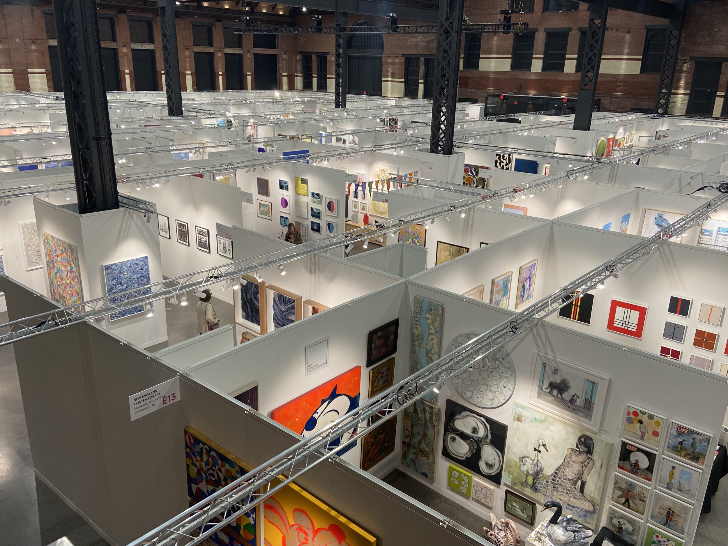 Interior view of an art exhibit with several walls filled with colorful paintings and artworks, in a large room with brick walls and steel support beams.