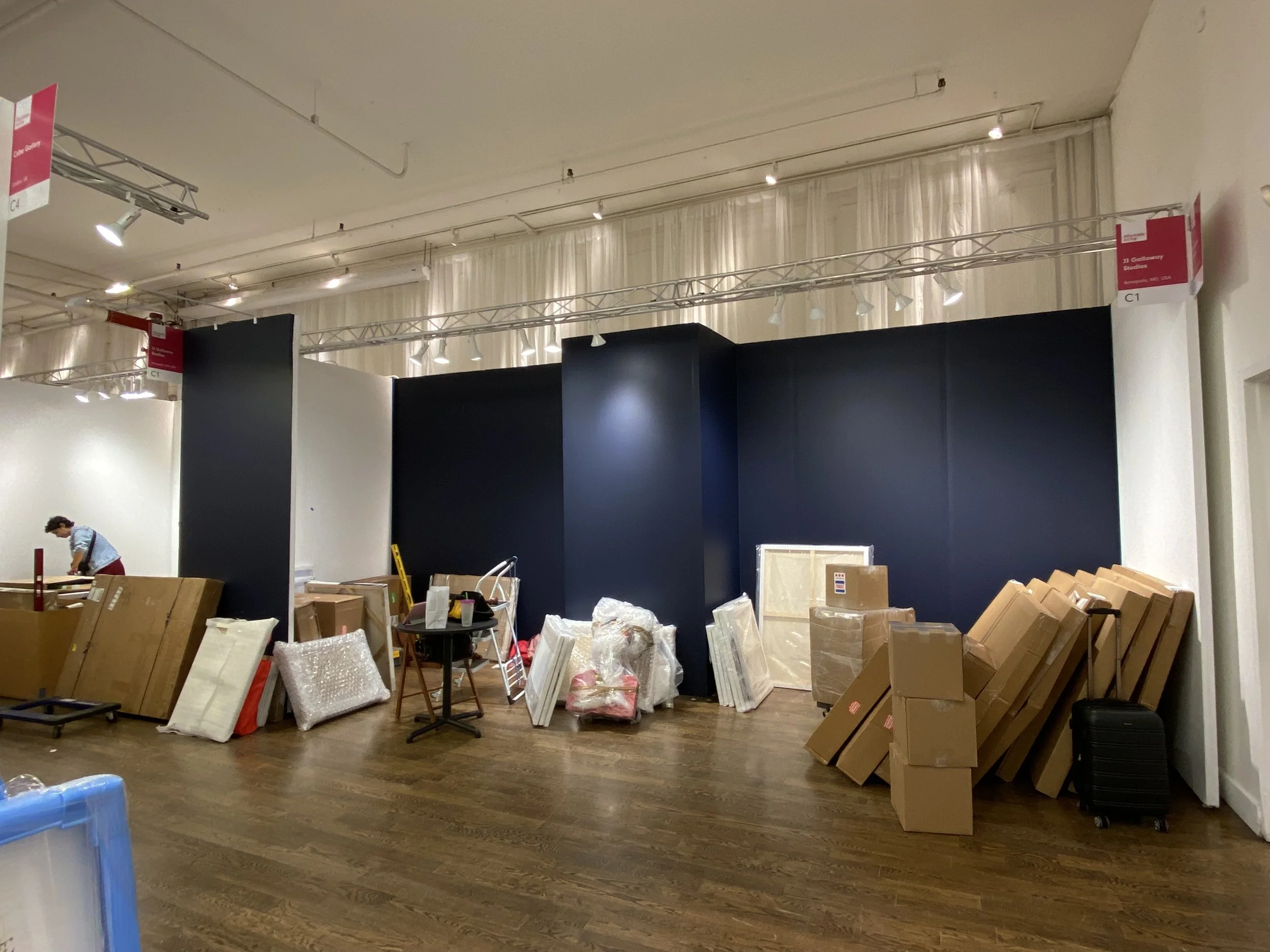 Interior of a room with various packages, boxes, and artwork supplies scattered across the floor, with black display walls and ceiling-mounted lights.