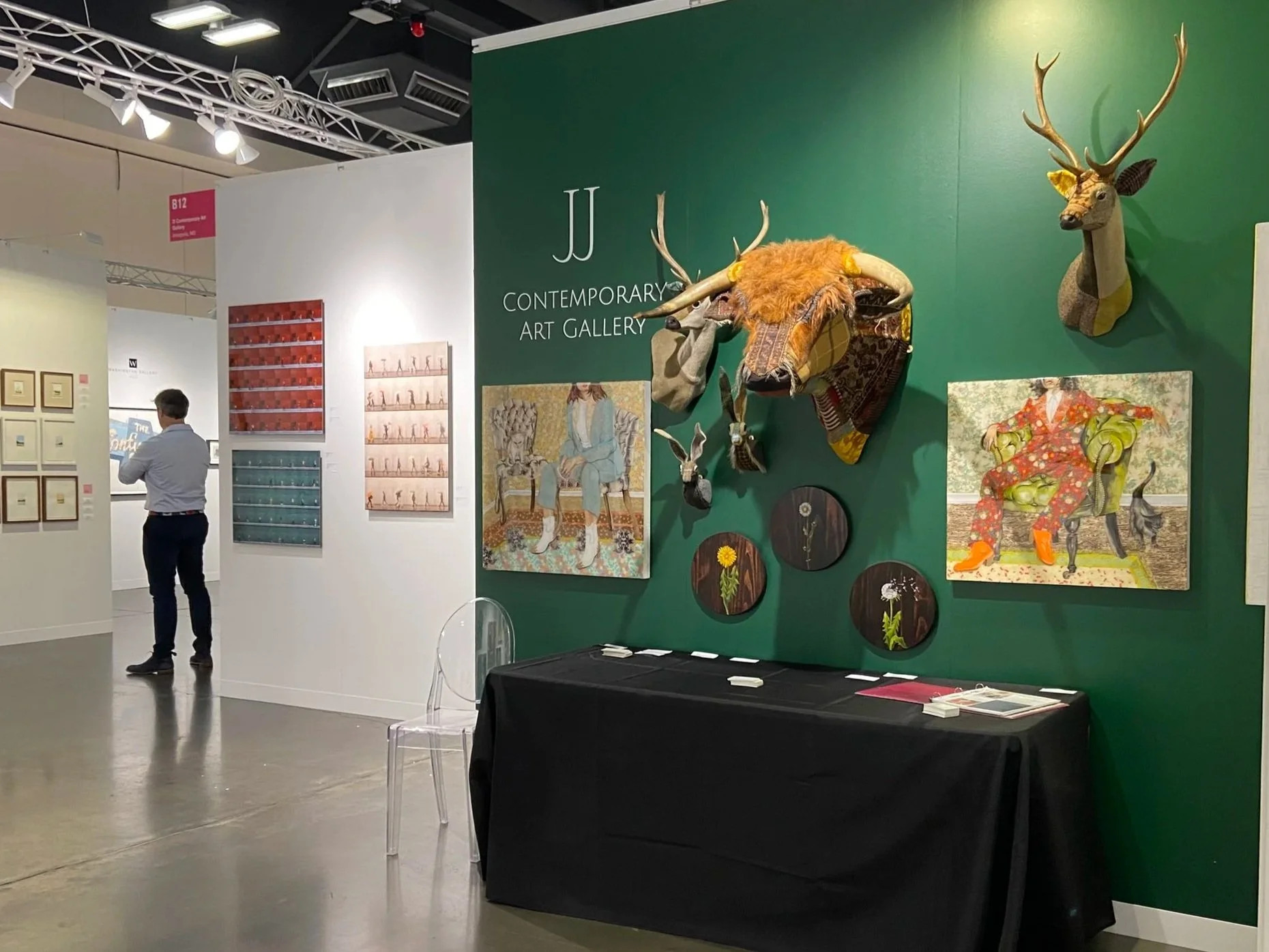An art gallery exhibit with a green wall displaying mounted animal sculptures, including a deer with large antlers and a head with a furry mane, alongside paintings of a woman sitting on a throne and a woman in a floral red dress. A person is looking at artwork on a white wall in the background.