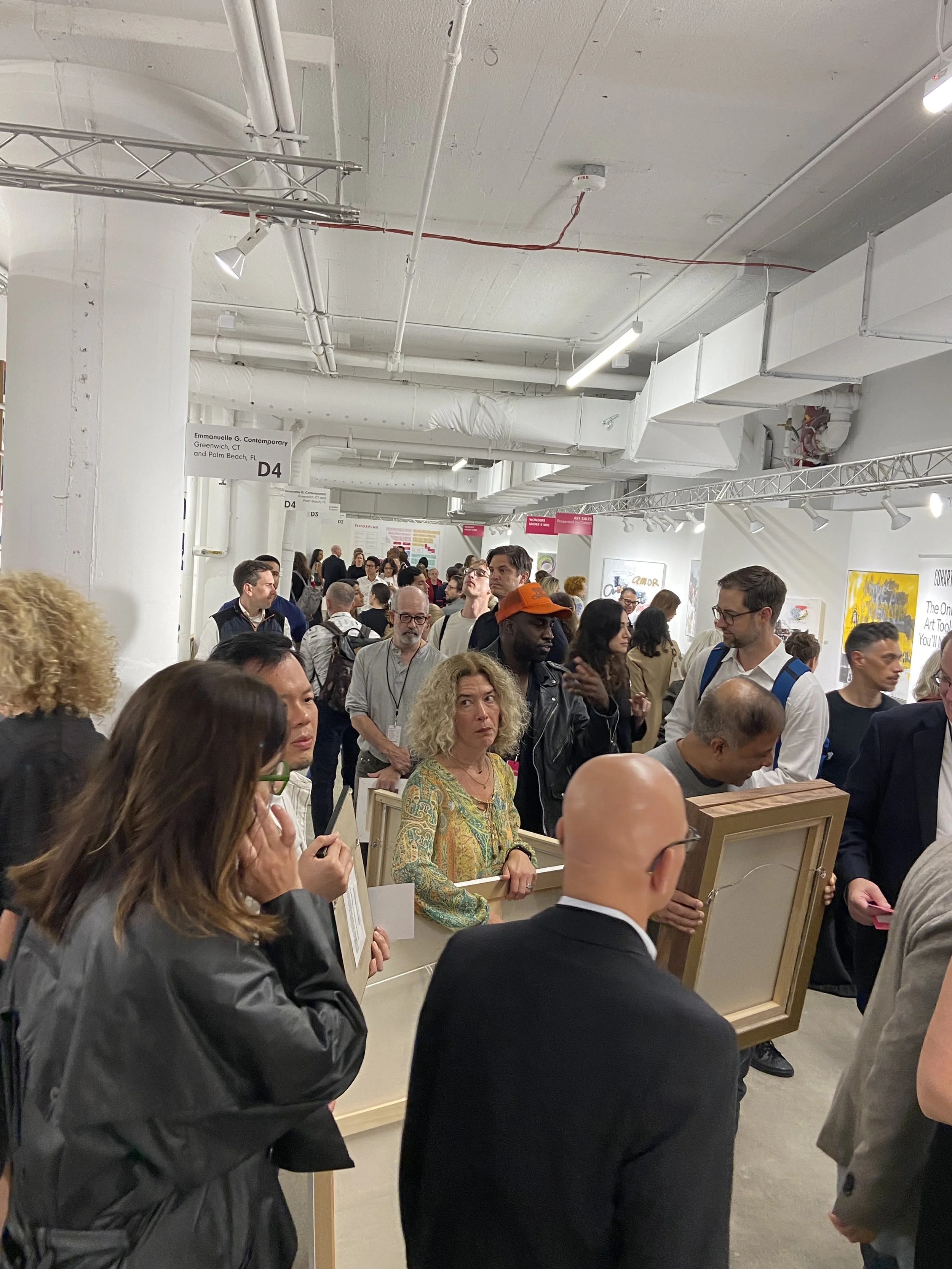 A crowded art exhibit with people viewing framed artworks displayed on tables. There are signs indicating different sections, and the space has white walls with commercial lighting and exposed ceiling pipes.