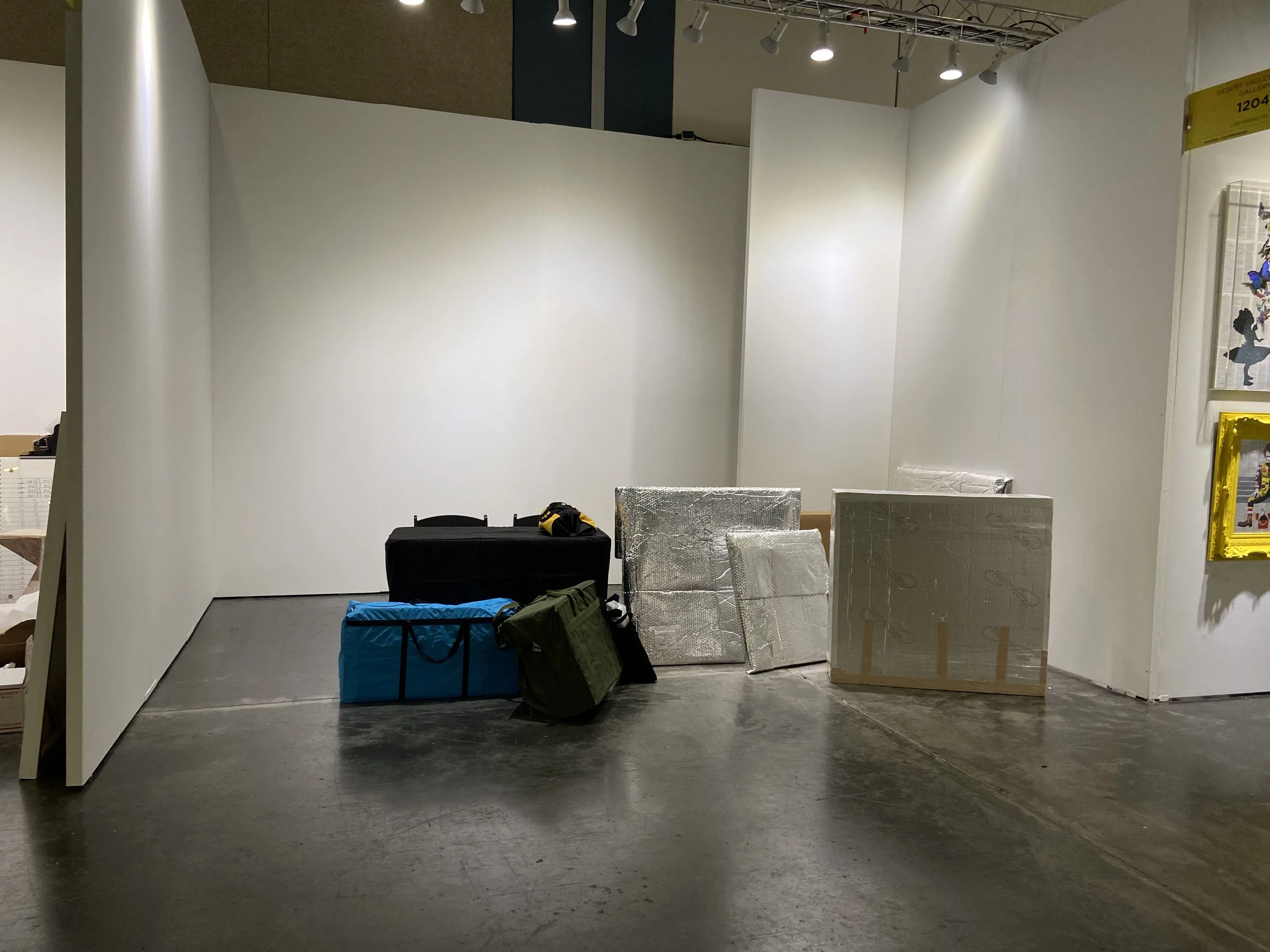 Empty exhibition booth with white walls, some packed boxes, and wrapped display items, set up in an indoor convention or trade show space.