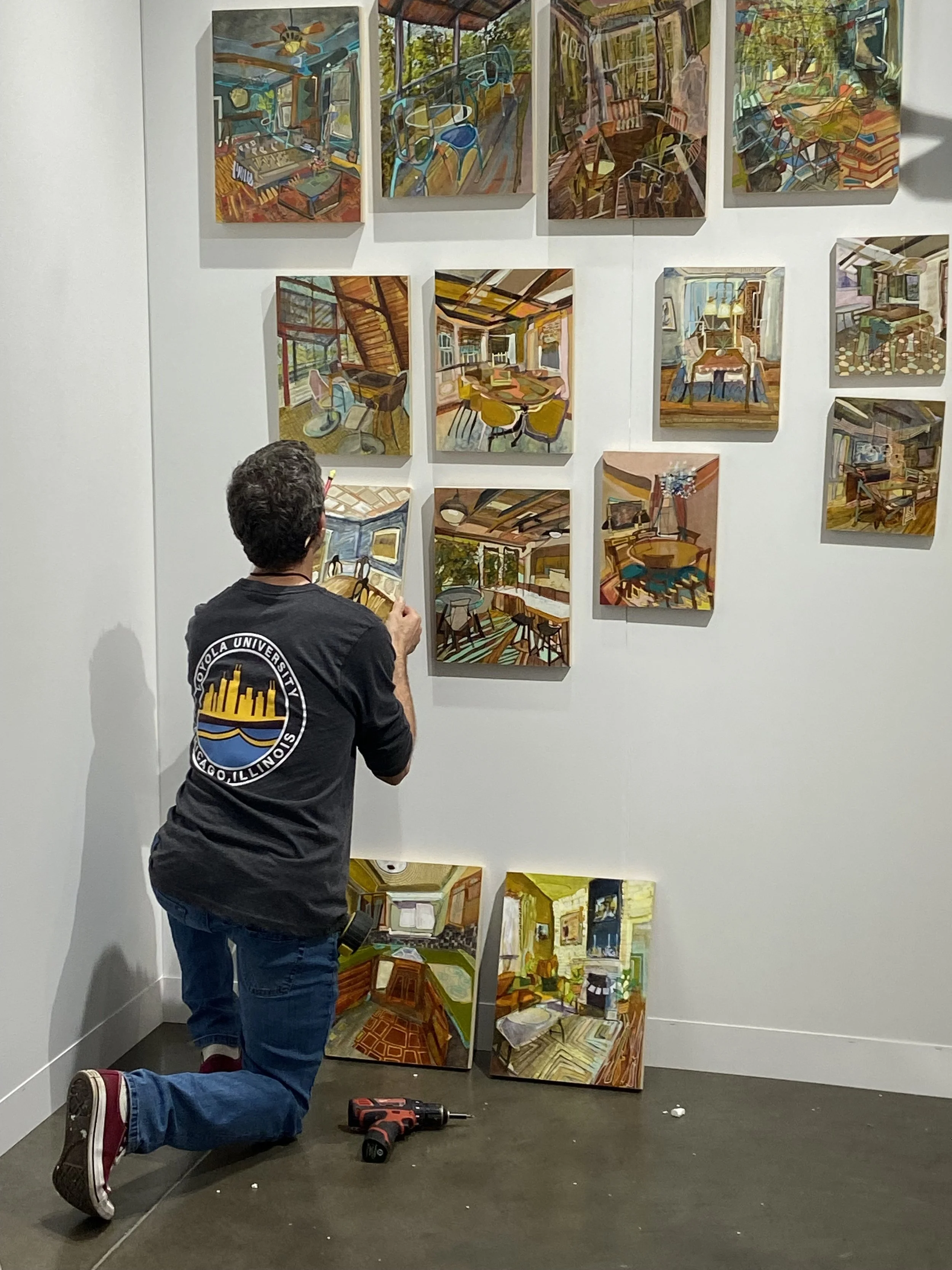 A person kneeling on the floor in front of a white wall with multiple colorful abstract paintings, some hanging on the wall and others stacking on the floor. The person is wearing a black shirt with a circular logo on the sleeve, blue jeans, and sneakers. Tools are on the floor nearby.