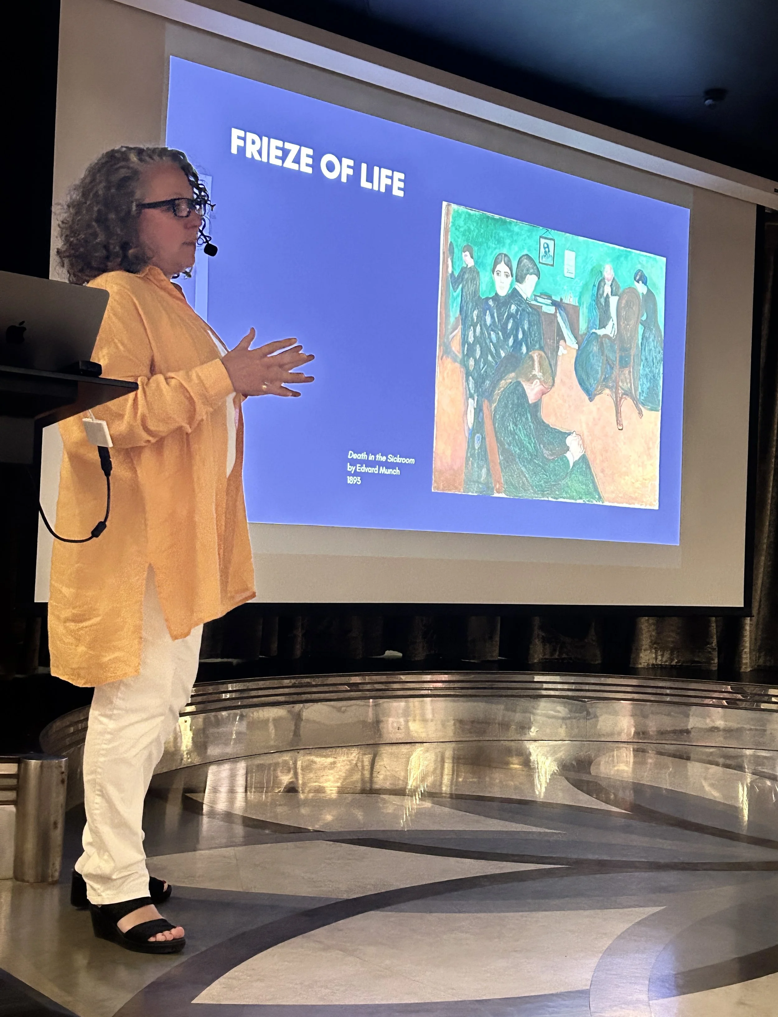 A woman with curly gray hair and glasses giving a presentation in front of a slide in a conference room. The slide shows a painting titled 'Death in the Sickroom' by Edvard Munch from 1893, with the words 'FRIEZE OF LIFE' at the top.