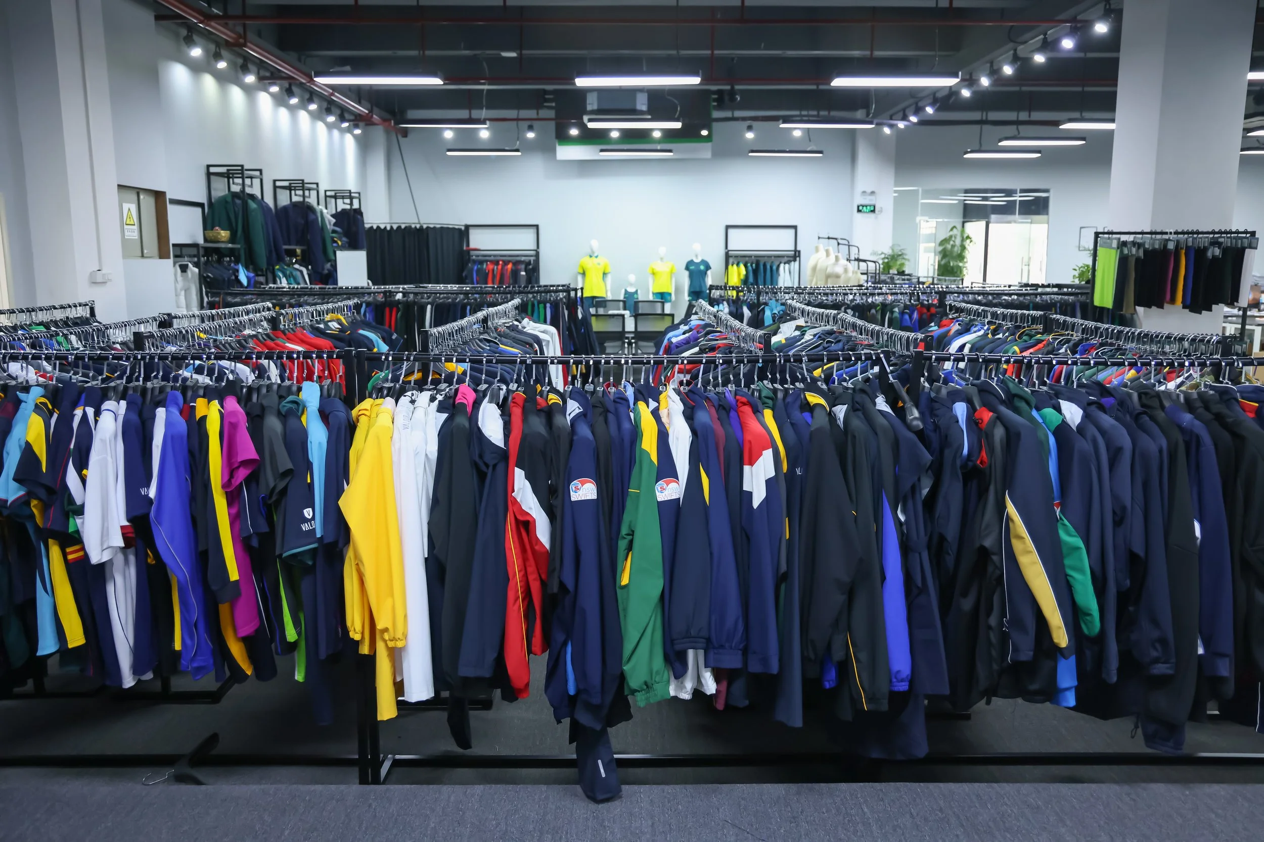 Indoor sportswear store with racks of colorful athletic shirts and jackets, mannequins dressed in sports uniforms in the background, lit by bright ceiling lights.