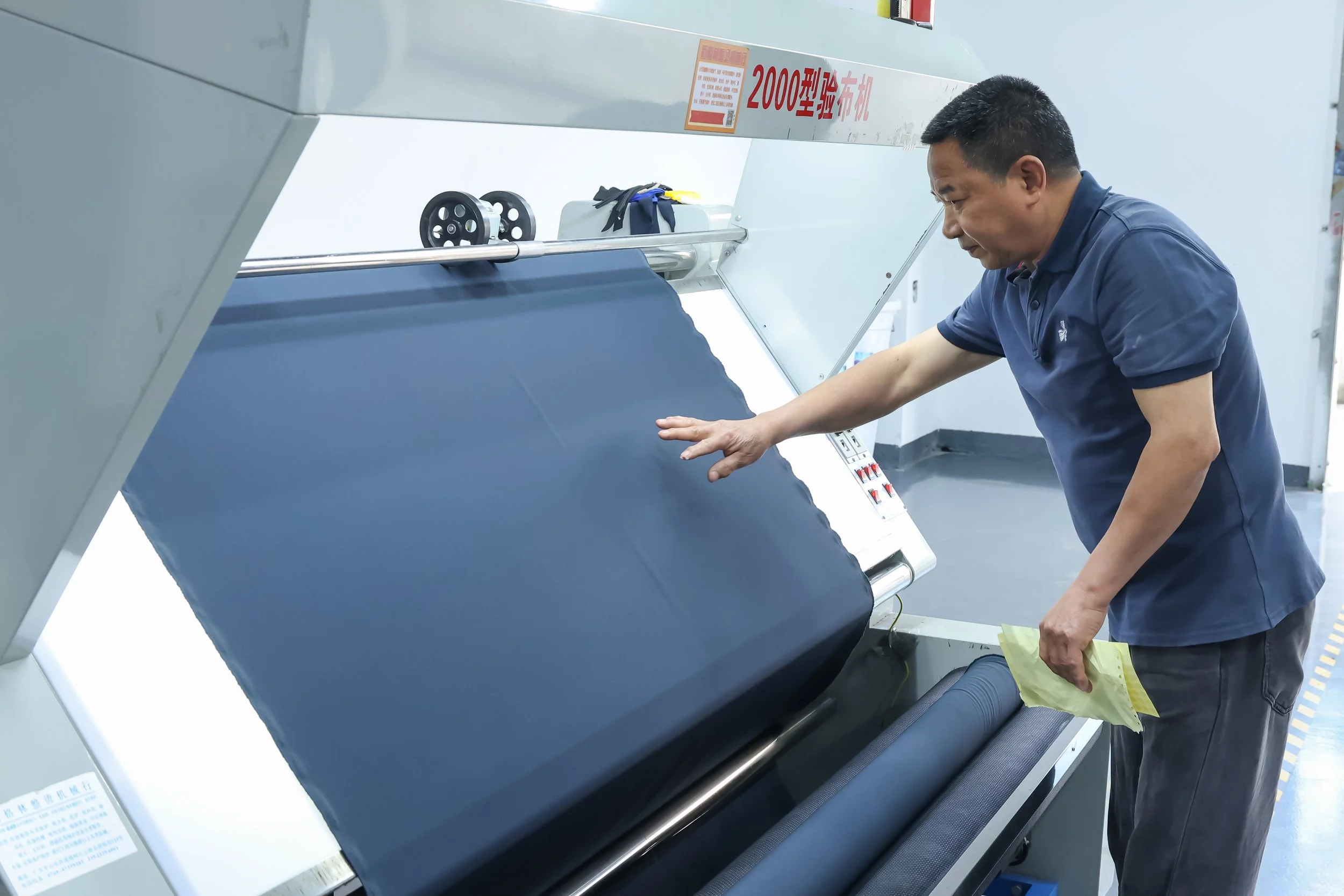 A man demonstrating the use of a large fabric cutting machine in a workshop or factory, wearing a blue polo shirt, and working on a roll of dark fabric.