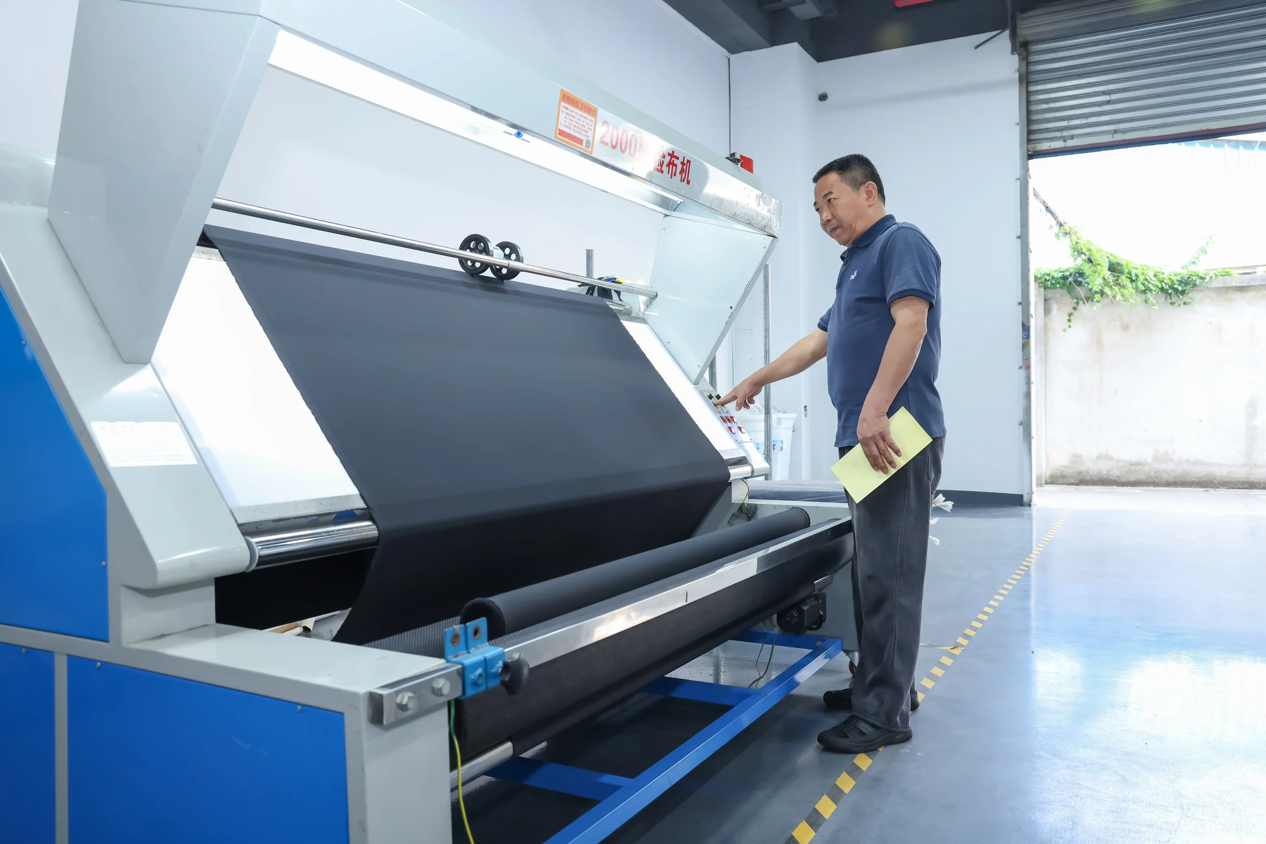 A man wearing a blue polo shirt and dark pants is standing next to a large industrial textile machine in a workshop or factory. He is holding a yellow piece of paper and appears to be operating or inspecting the machine, which has Chinese characters and a red sign with white text on it. The workspace has a concrete floor with yellow dashed lines, and an open garage door showing some greenery outside.