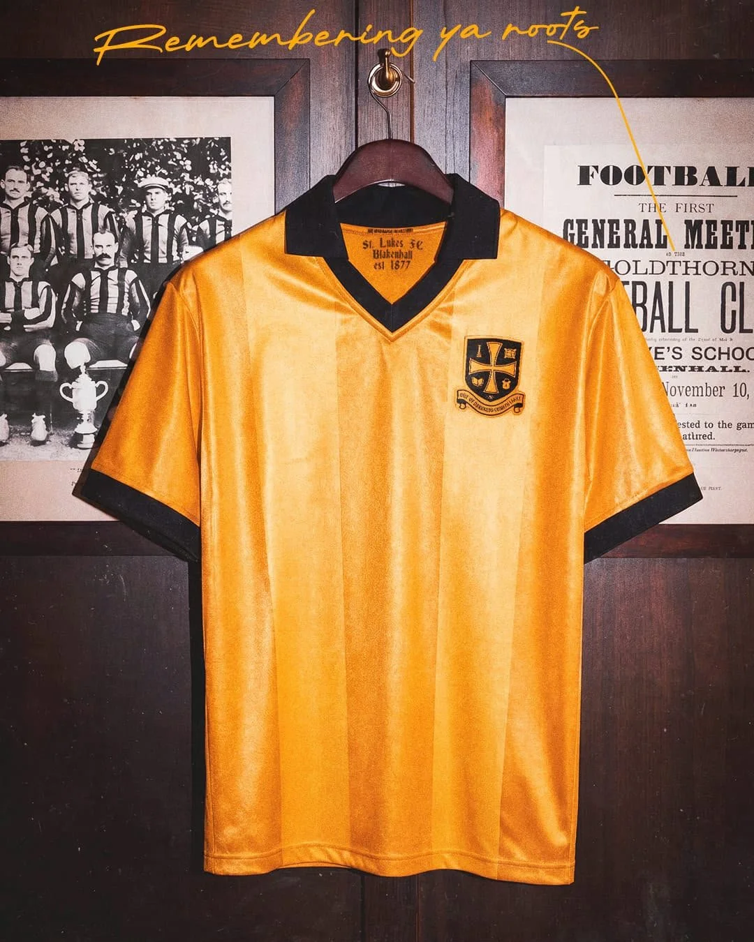 Yellow soccer jersey with black collar and sleeve trim, embroidered crest on the chest, hung on a wooden wall with vintage sports photographs and posters.