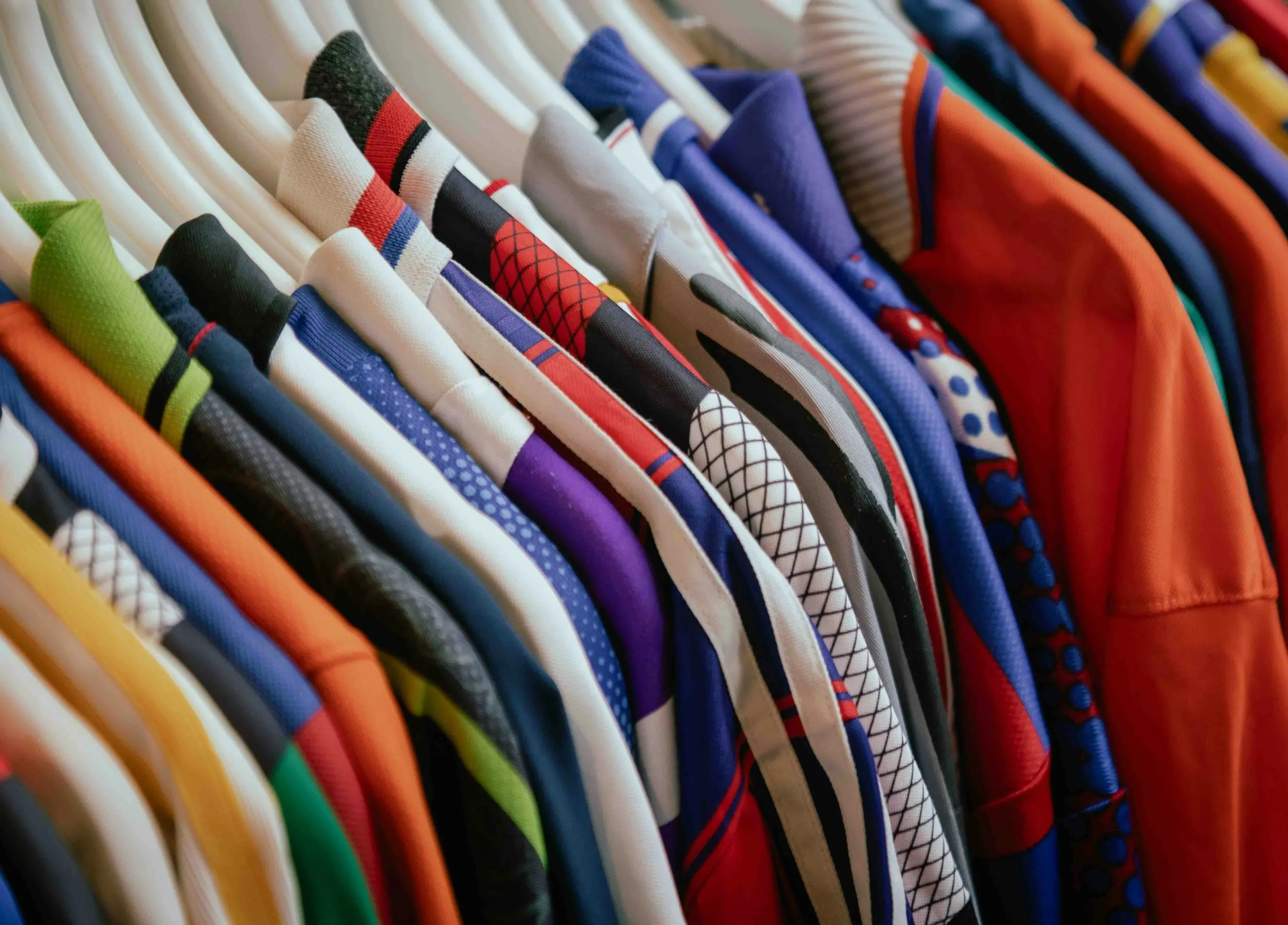 A row of colorful sports jerseys hanging on white hangers. The jerseys feature various patterns, colors, and designs, including stripes, polka dots, and mesh.