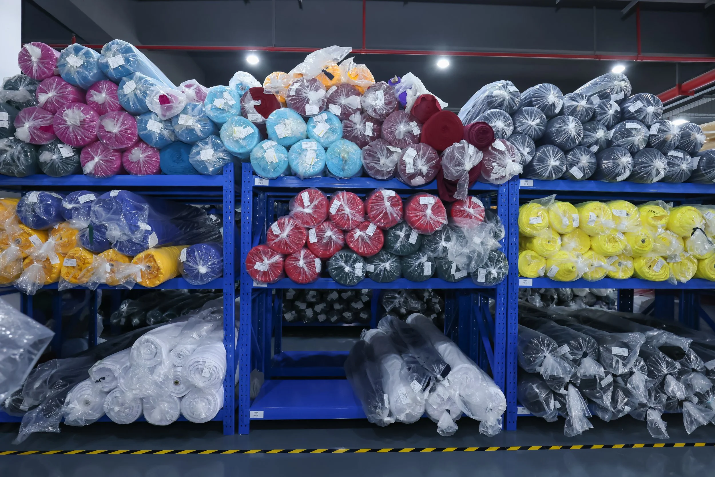 Shelves filled with colorful rolls of fabric or material in a warehouse store, organized by color including pink, blue, purple, yellow, black, and red.