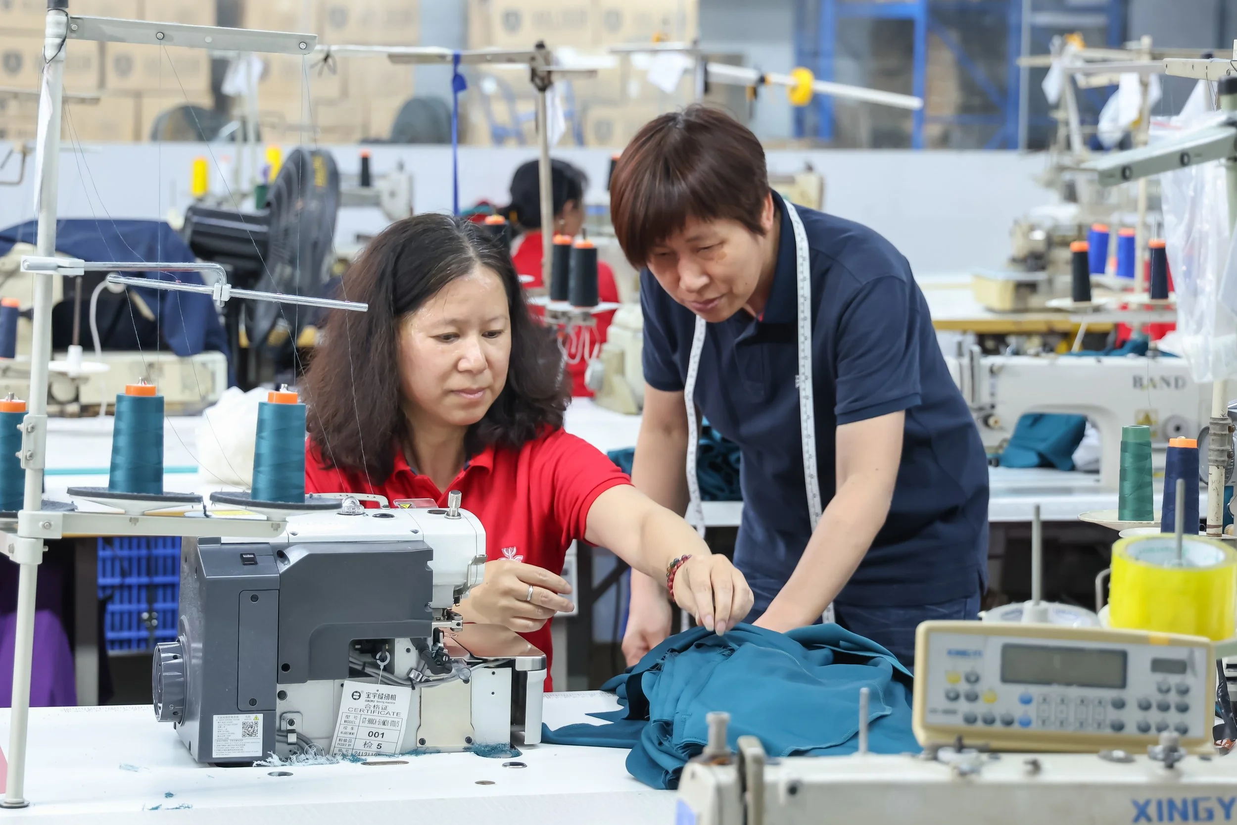 Two women working together on a sewing machine in a factory, surrounded by spools of thread and sewing equipment, with other workers in the background.