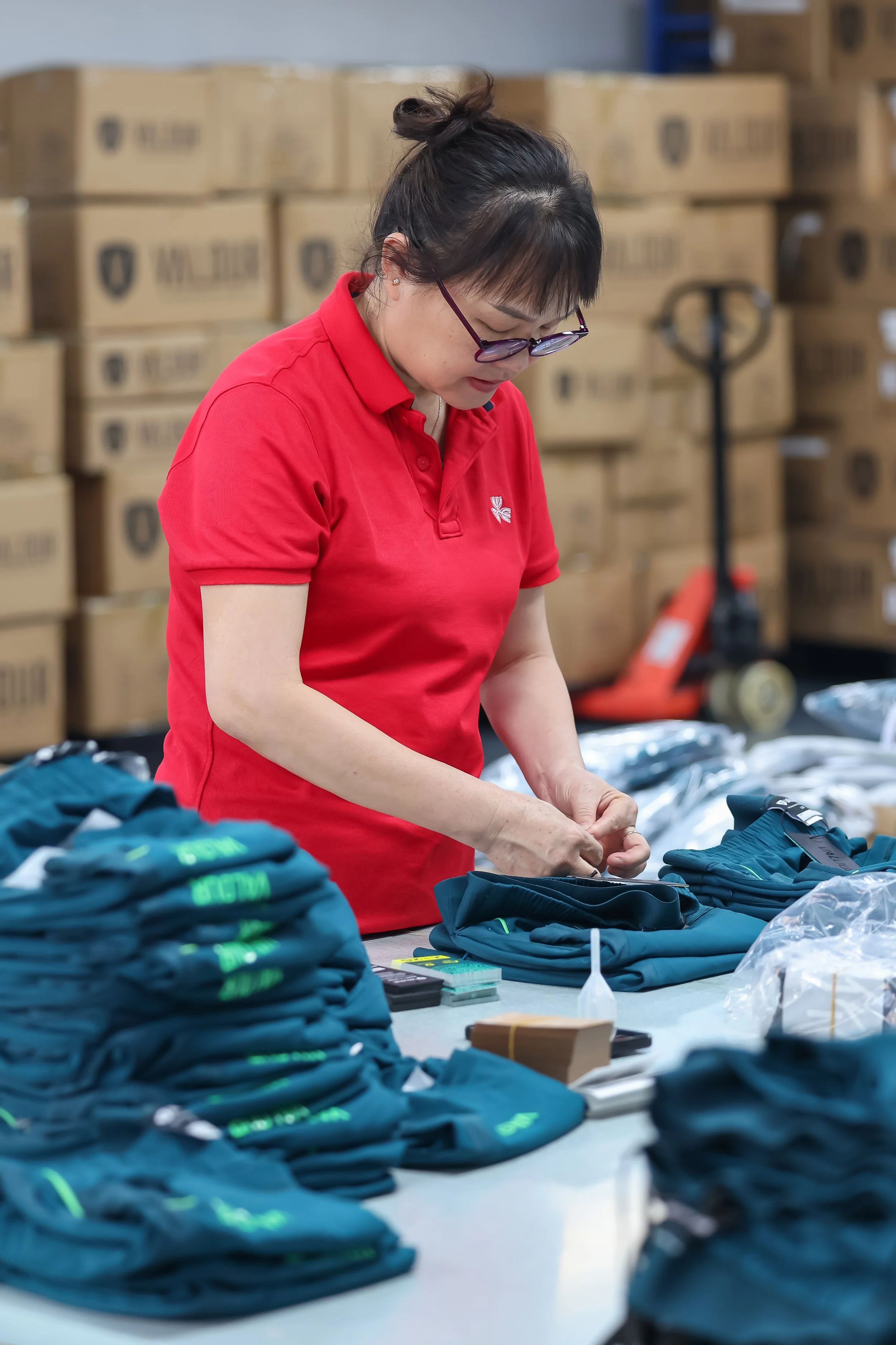 A woman in a red polo shirt and glasses gathers items at a table surrounded by stacks of blue and black clothing or gear, with boxes in the background in a warehouse or storage facility.