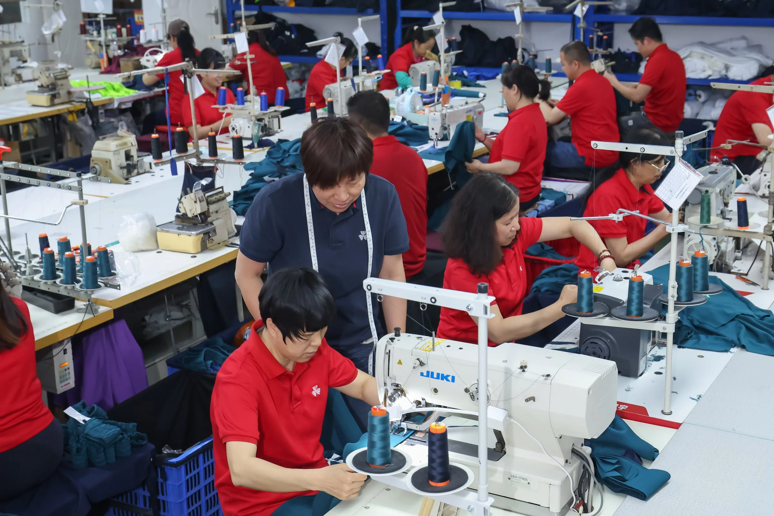 Group of workers assembling garments at sewing machines in a garment factory. The workers are wearing red shirts.