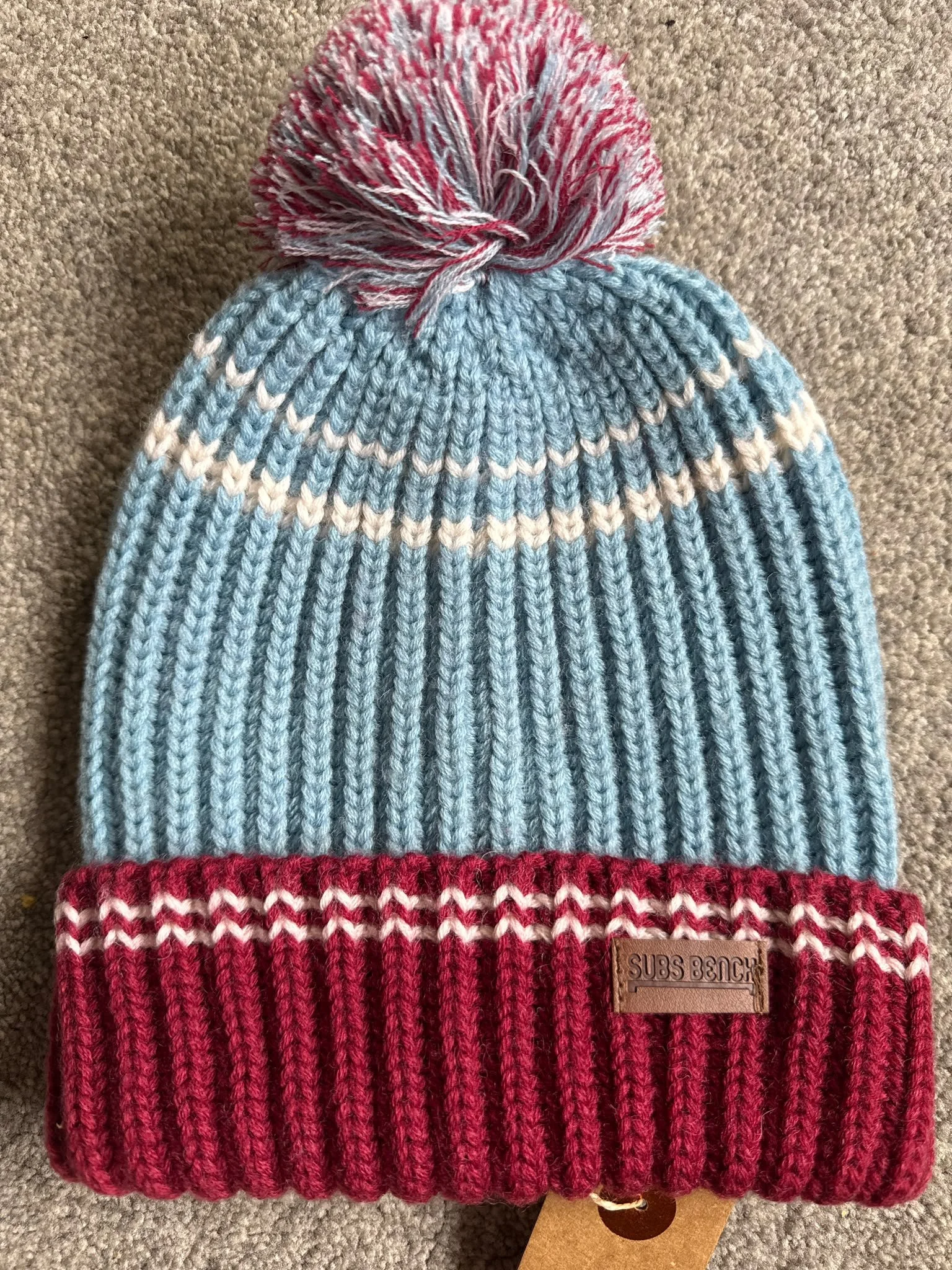 A knitted beanie hat with a pom-pom on top, featuring light blue, white, and red colors, with a leather tag that reads 'SUBS BENCH' at the bottom.