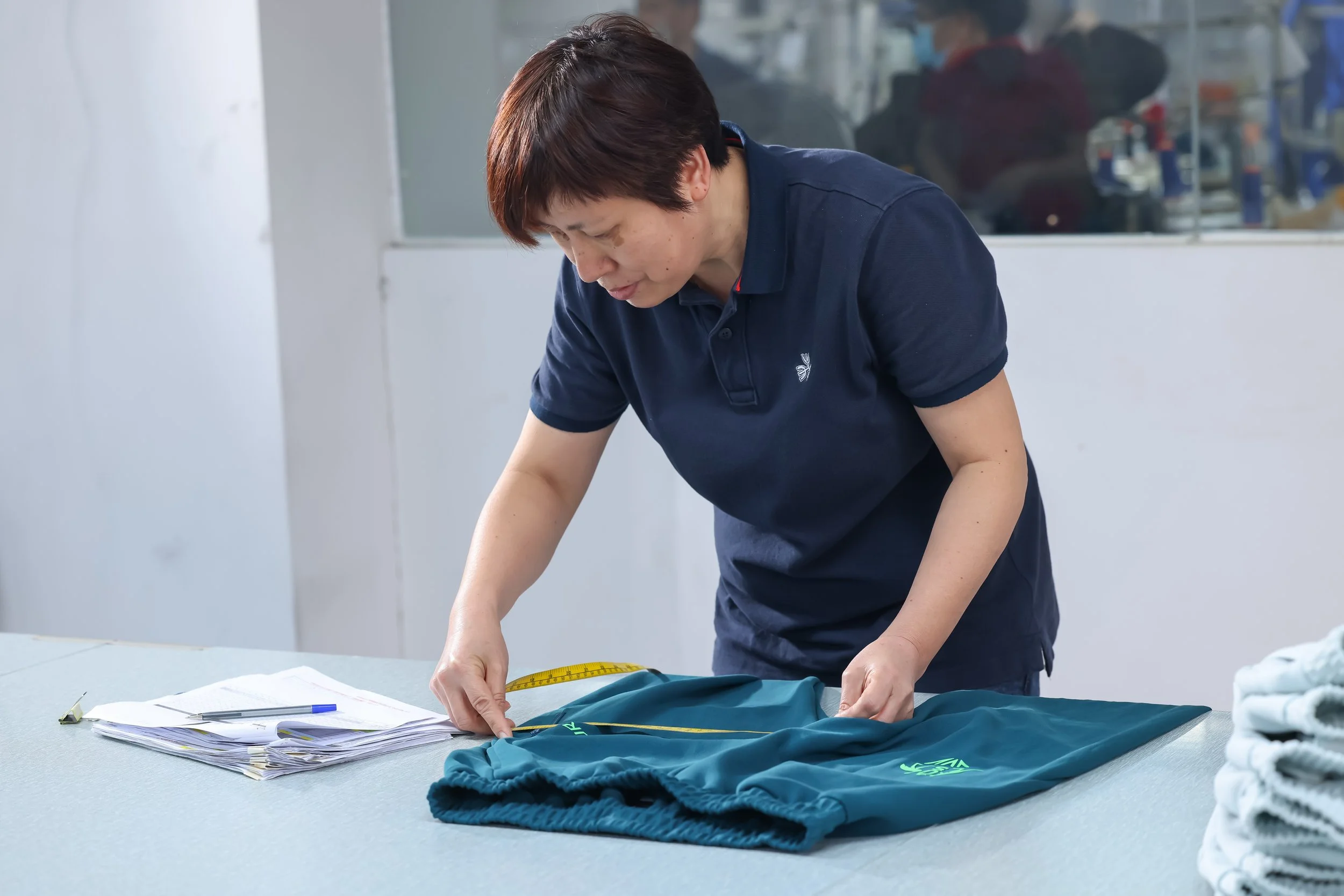 A woman is working with clothing and papers on a table, measuring a piece of clothing with a tape measure.