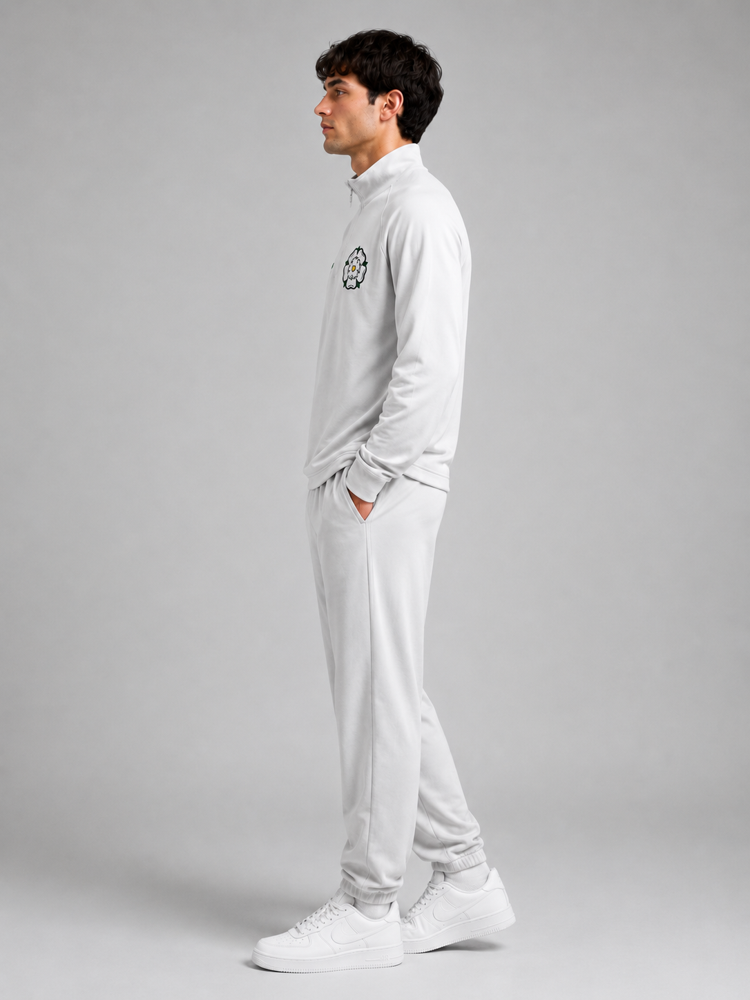 Side view of a young man with dark, curly hair wearing an all-white tracksuit, standing against a gray background.