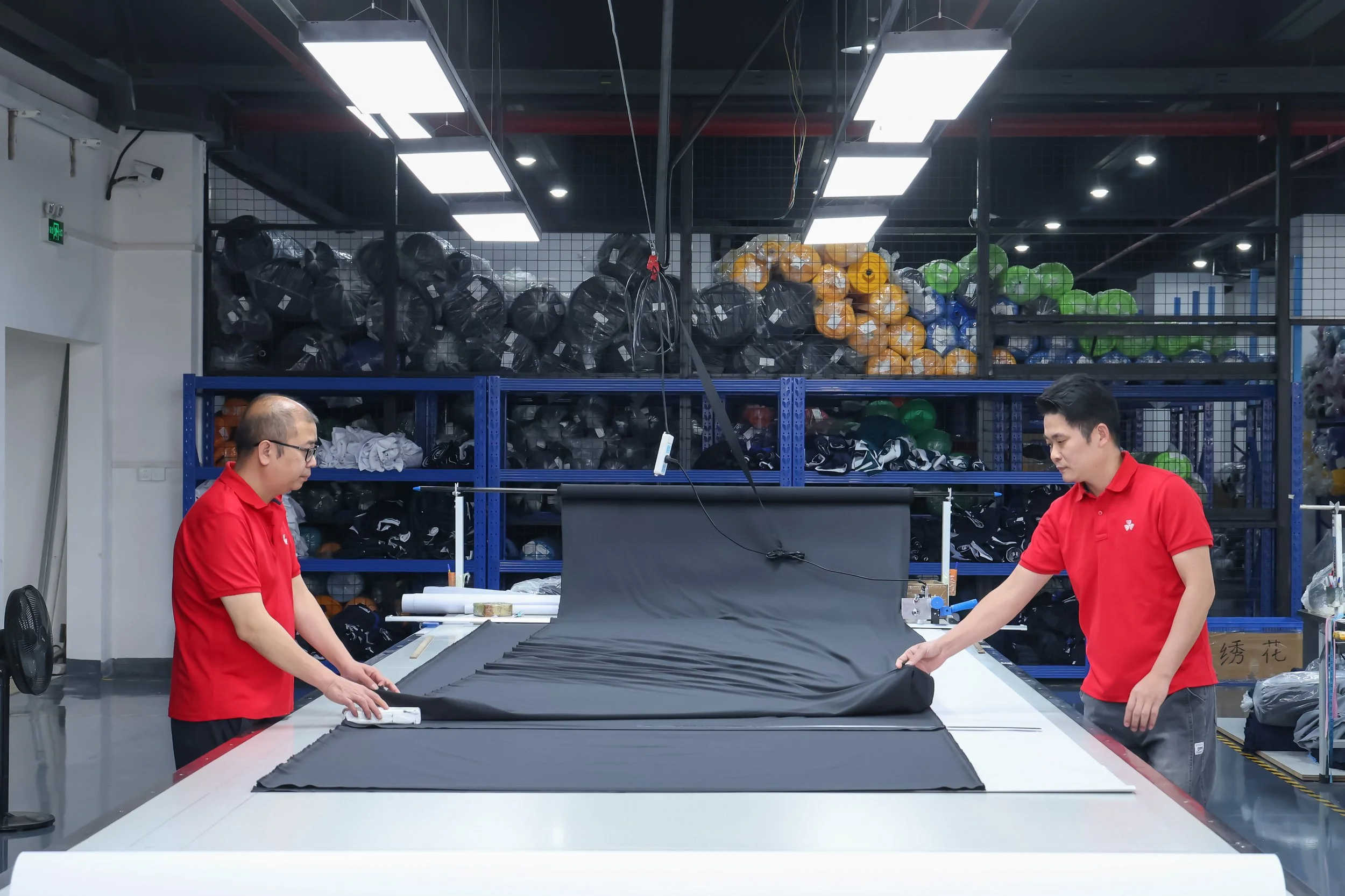 Two workers in red shirts are working on a large black fabric in a well-lit industrial workshop filled with sports equipment, with shelves holding black, orange, green, and blue balls in the background.