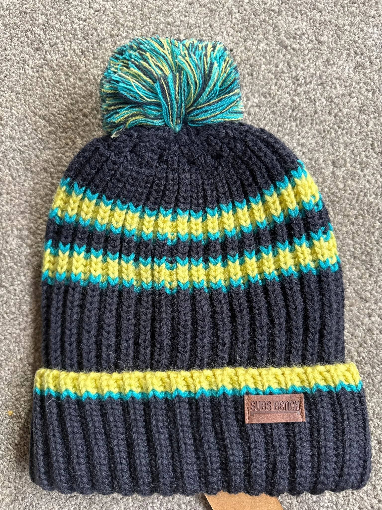 Close-up of a knitted beanie hat with a pom-pom on top, featuring black, yellow, and blue stripes, and a leather patch that says 'SUBS BENCH' on a gray carpet.