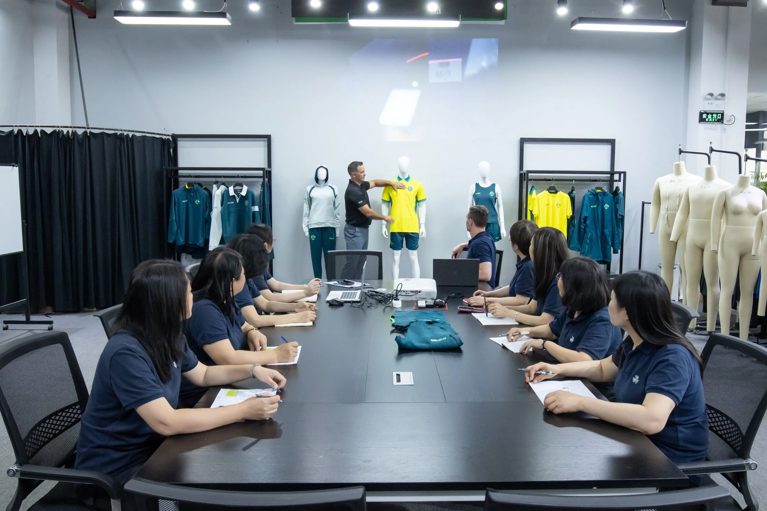A group of people seated around a large conference table, attending a presentation in a room with sportswear on display mannequins and racks.