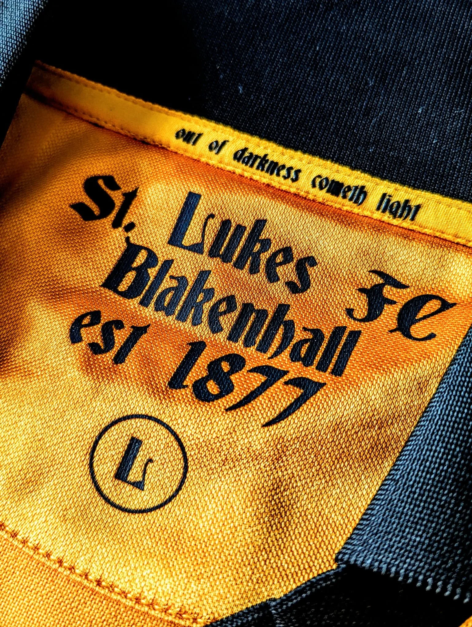 Close-up image of a yellow fabric label with black text, stitched onto a garment, showing the words 'Lufkes' and 'Blakes' and some additional text, with part of a black fabric and blue denim visible.