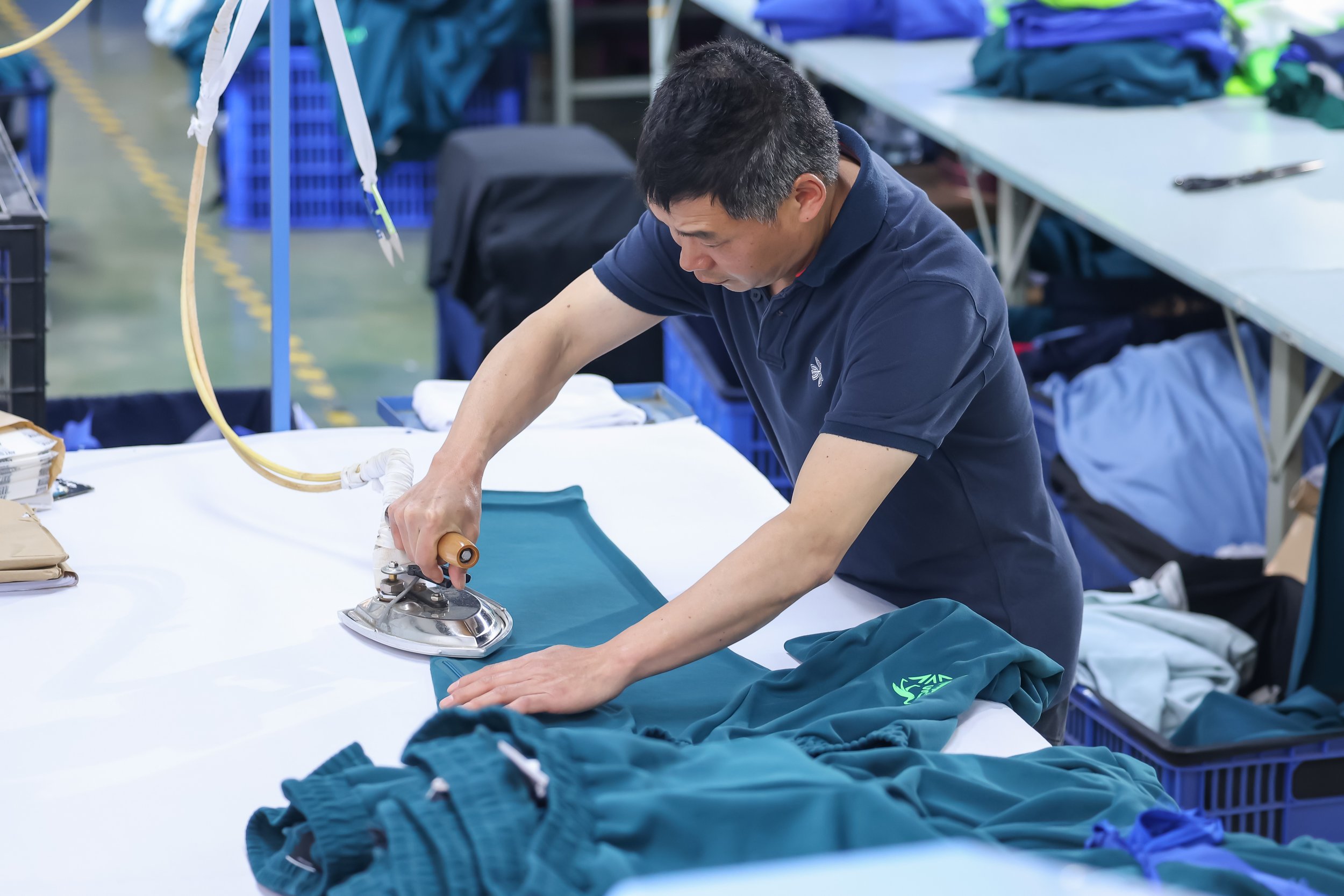 A person is ironing fabric on a table in a workspace, surrounded by folded clothes and textile supplies.