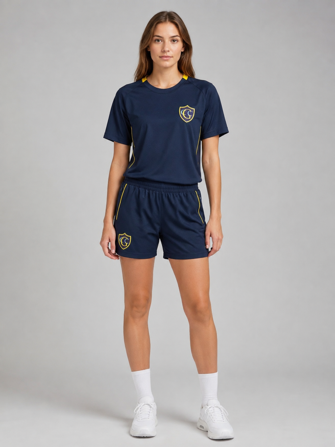 A woman wearing a navy blue sports uniform consisting of a T-shirt and shorts with yellow accents and logos, white sneakers, and white socks, standing against a plain gray background.