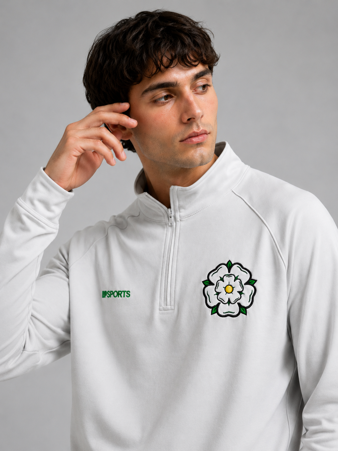 A young man with dark curly hair wearing a white sports jacket with a black and white embroidered flower logo on the chest, and the word 'UPSPORTS' in green on the left side, touching his ear with his right hand, against a plain gray background.