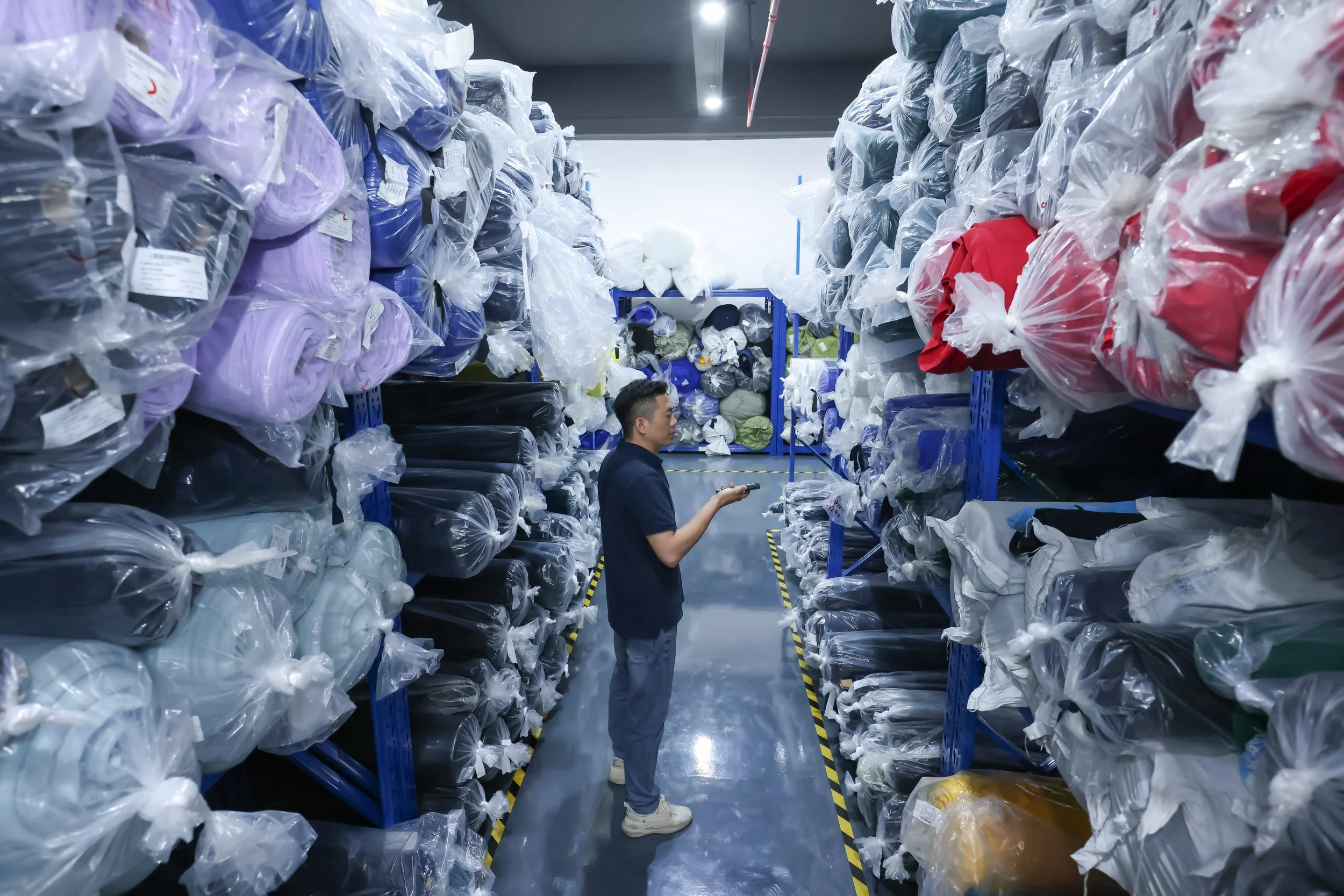 A person standing in a storage aisle surrounded by shelves filled with plastic bags of clothing in a warehouse.