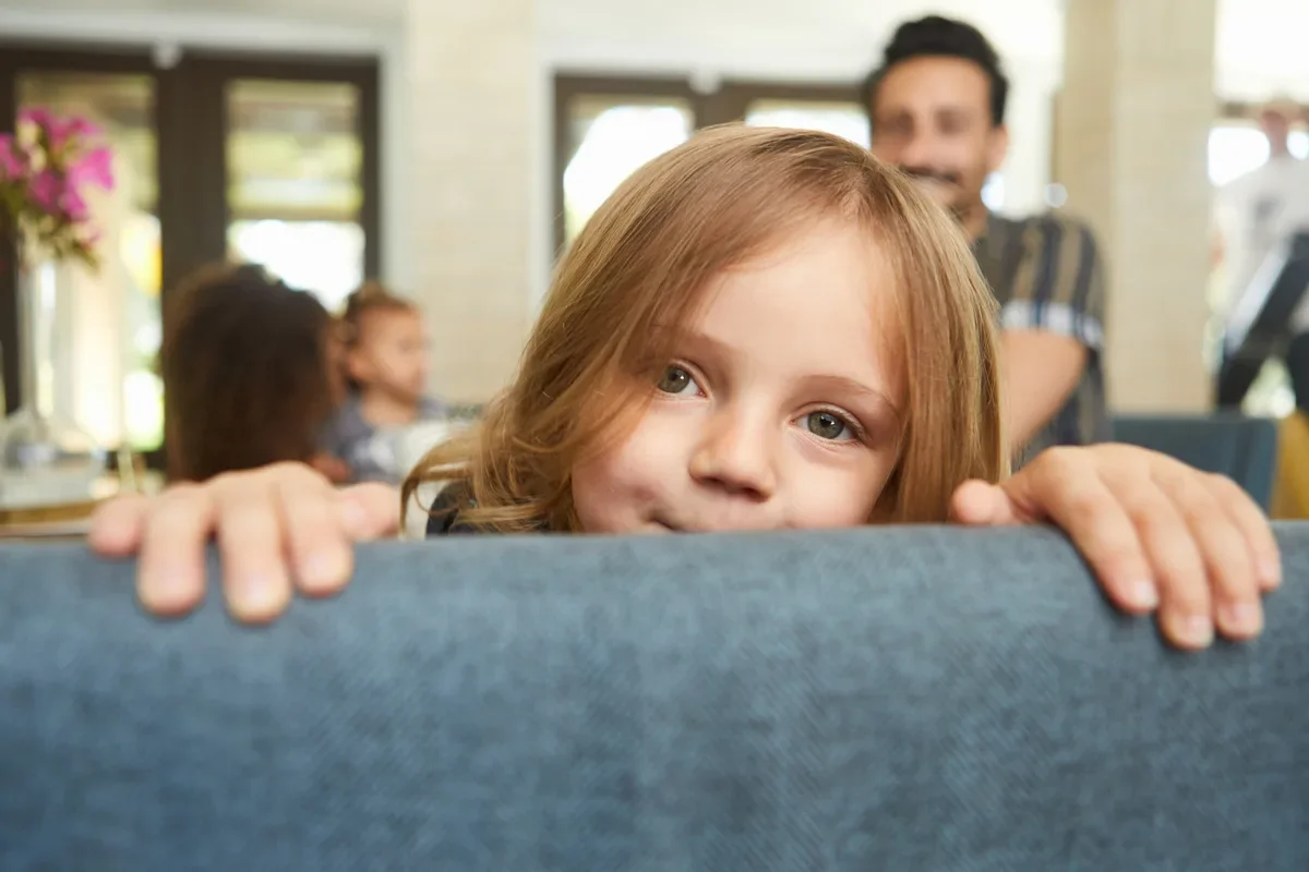 Young child with reddish hair peeking over couch cushion in bright modern home setting