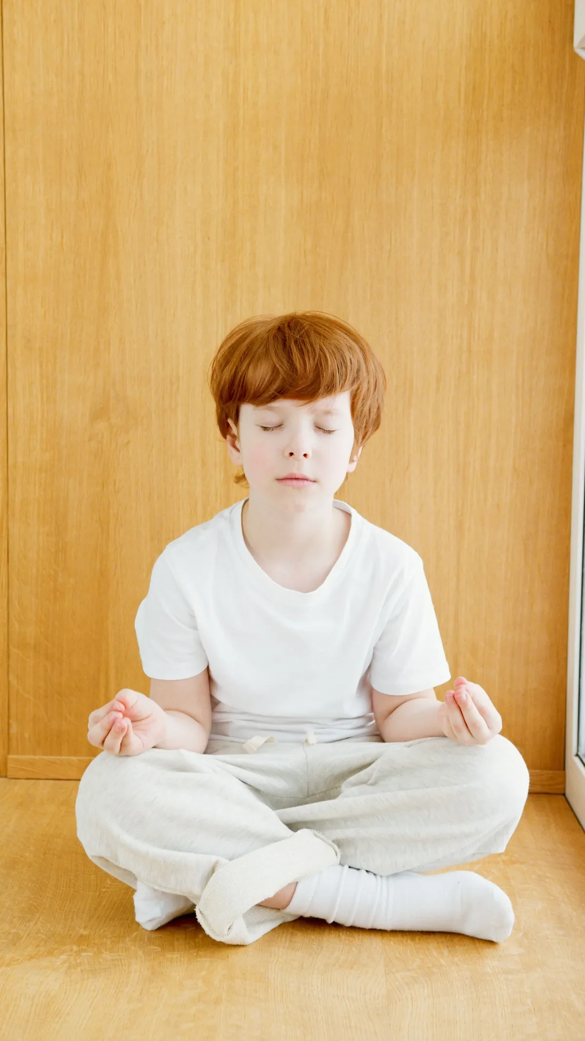Redhead child in white clothing seated cross-legged in calm meditative pose on wooden floor