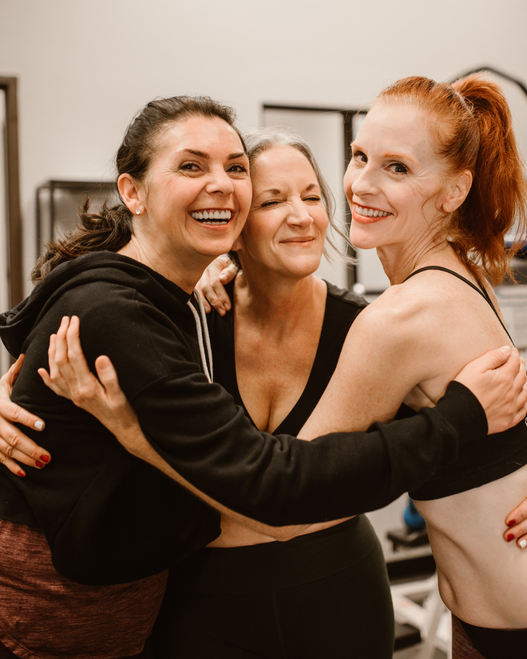 Três mulheres sorrindo e se abraçando, em um ambiente interno.