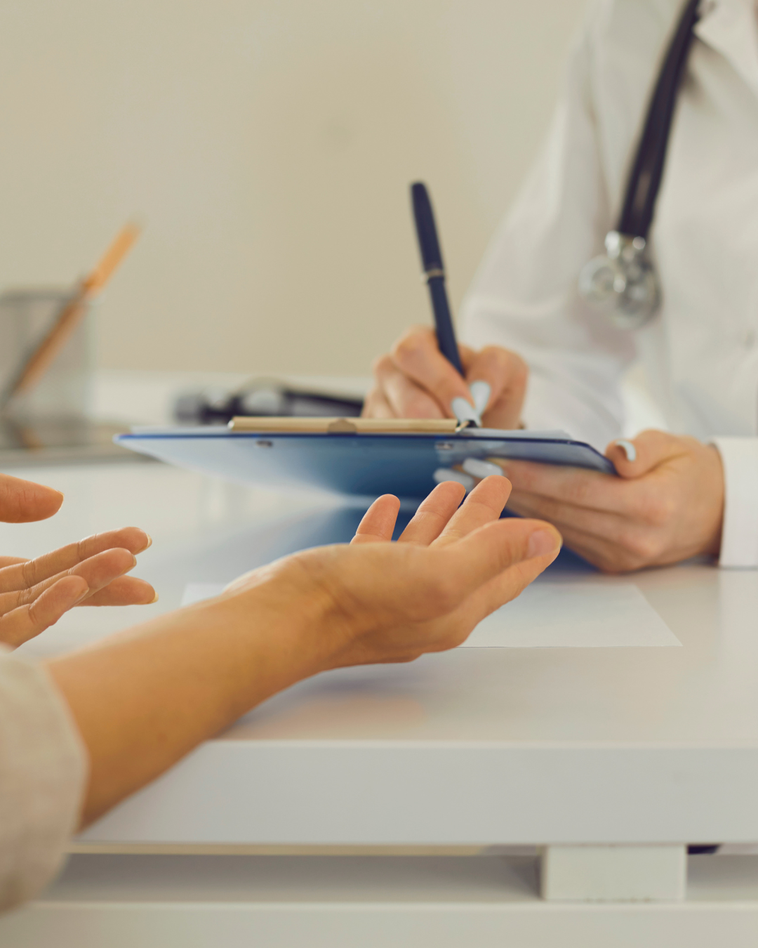 Uma pessoa, possivelmente paciente, estende a mão em direção a uma profissional de saúde, que está escrevendo em um prontuário ou ficha, em um ambiente de consulta médica.