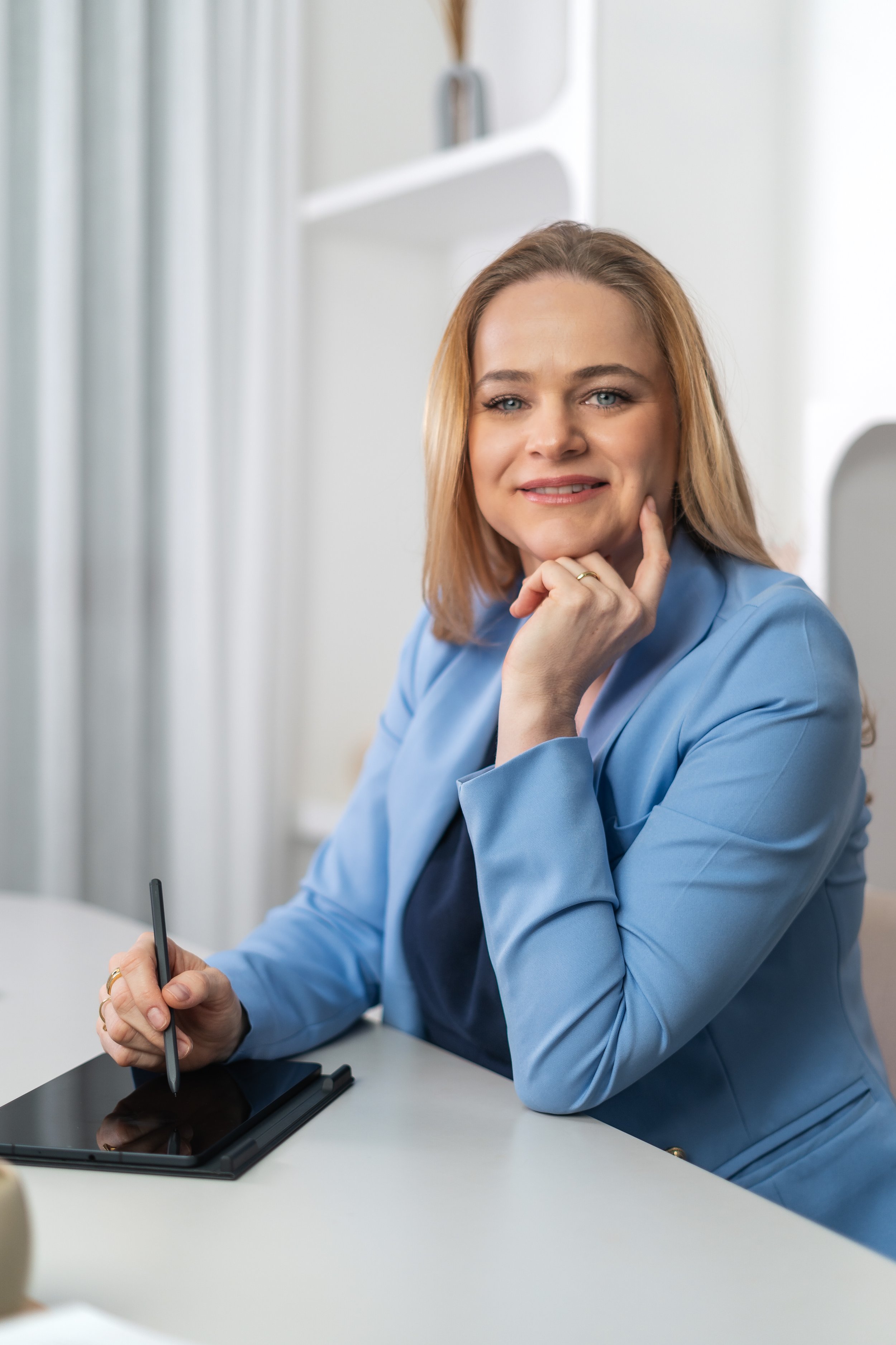 Médica Ginecologista em Florianópolis , usando blazer azul, sentada em uma mesa, segurando uma caneta com um tablet de desenho digital, com expressão amigável, em ambiente de escritório claro.