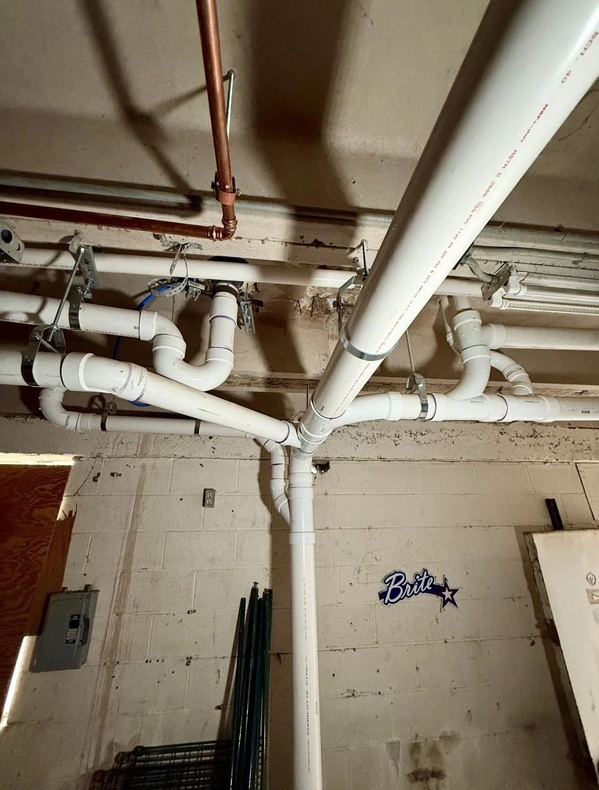 View of plumbing pipes and fittings mounted on a basement ceiling, including white PVC pipes, a copper pipe, and various connectors and brackets.