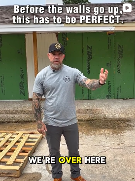 A man with a beard, wearing a gray T-shirt and a black cap, standing in front of a green construction wall, giving a thumbs up, with text overlay saying 'Before the walls go up, this has to be PERFECT.' and 'WE'RE OVER HERE'.