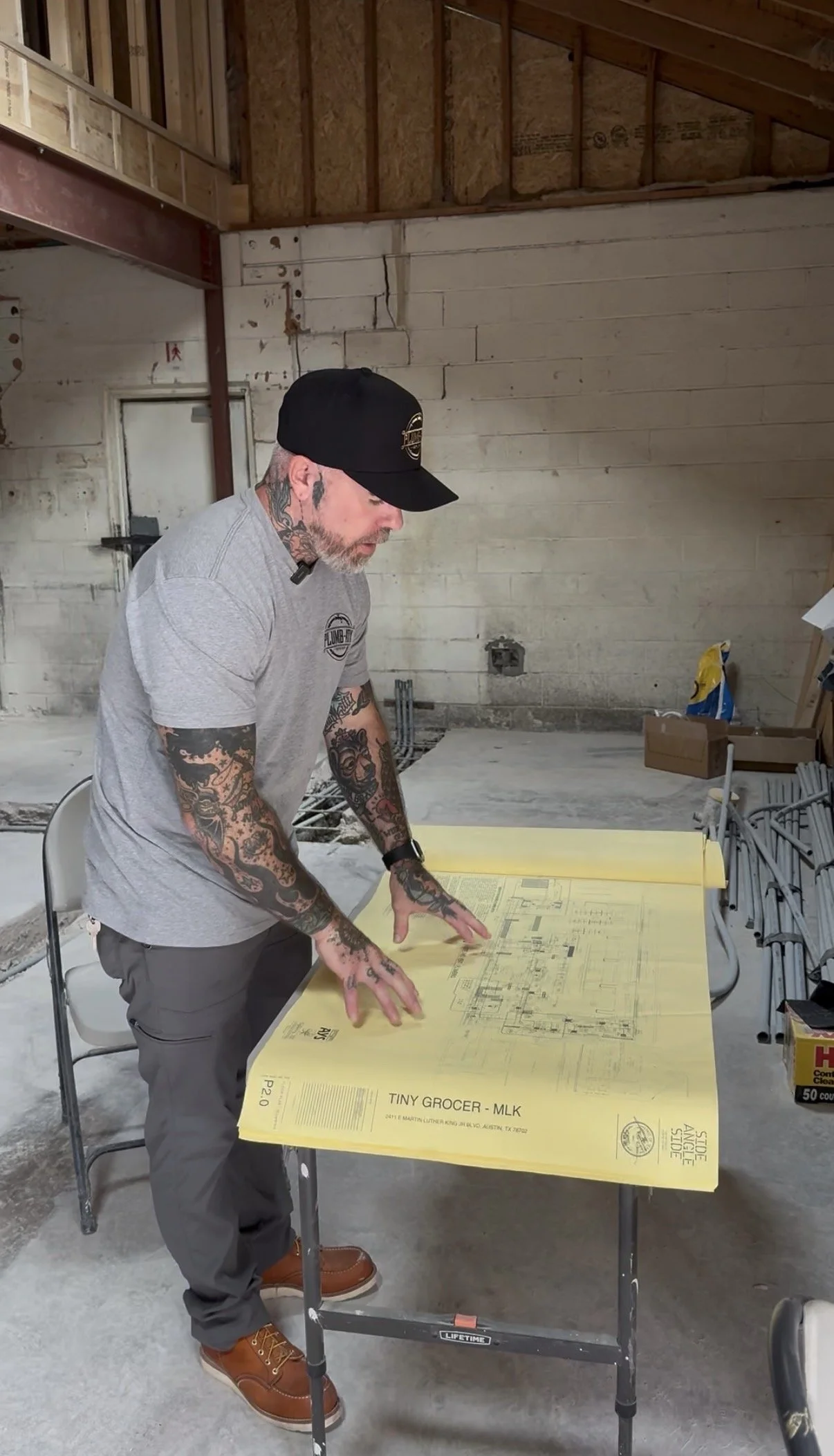A man with tattoos, wearing a black cap, gray T-shirt, and gray pants, is standing at a table looking at architectural blueprints spread out on the table in an unfinished building with exposed wooden framing and concrete walls.