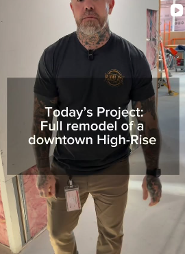 A man with tattoos wearing a black shirt with a logo, standing in an indoor construction area with tools and equipment in the background, with overlay text about a downtown high-rise remodel project.
