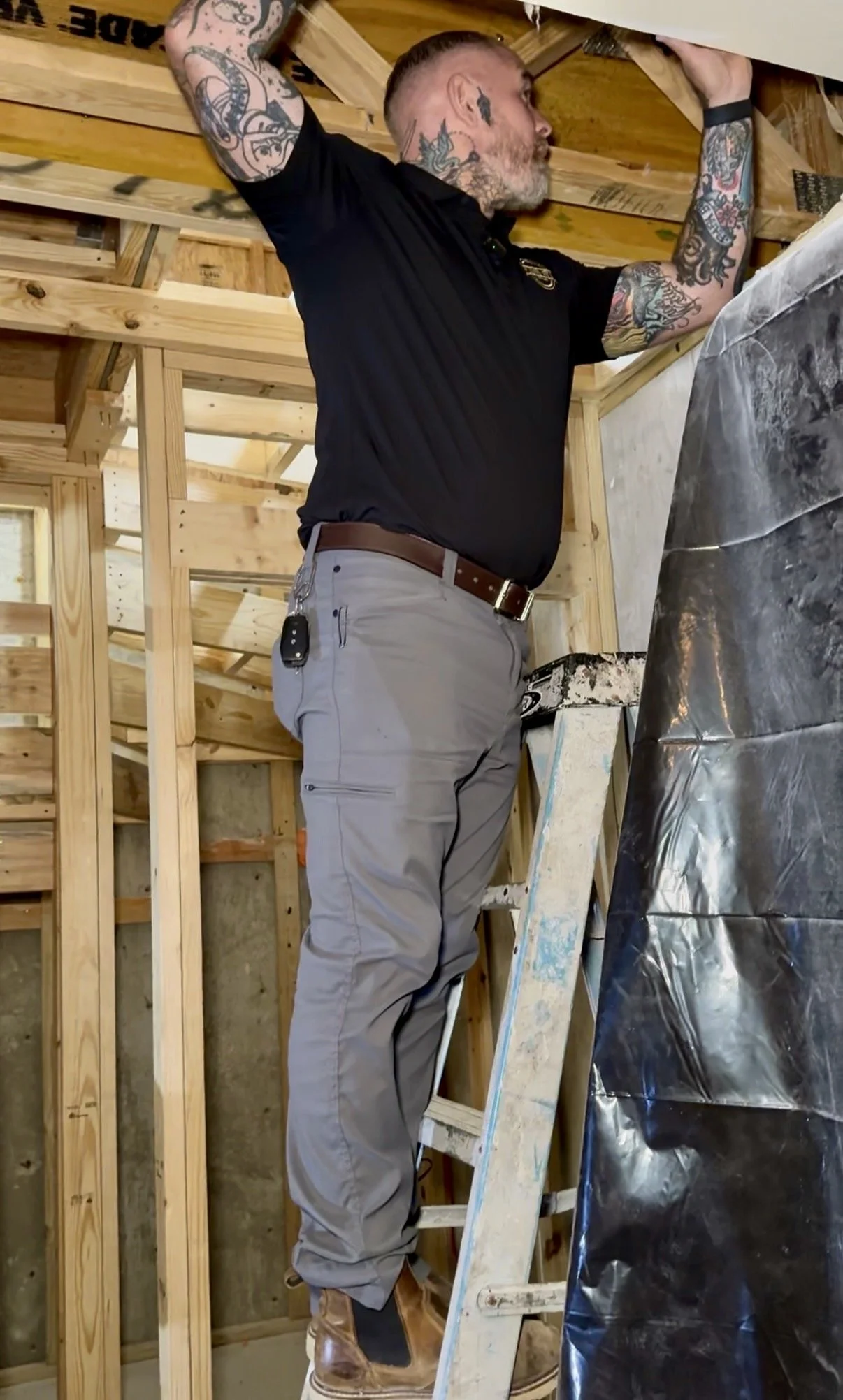 A man with tattoos on his arms and neck, wearing a black shirt, khaki pants, and brown work boots, is standing on a ladder working on the ceiling of a building under construction. The building has exposed wooden framing and insulation materials.