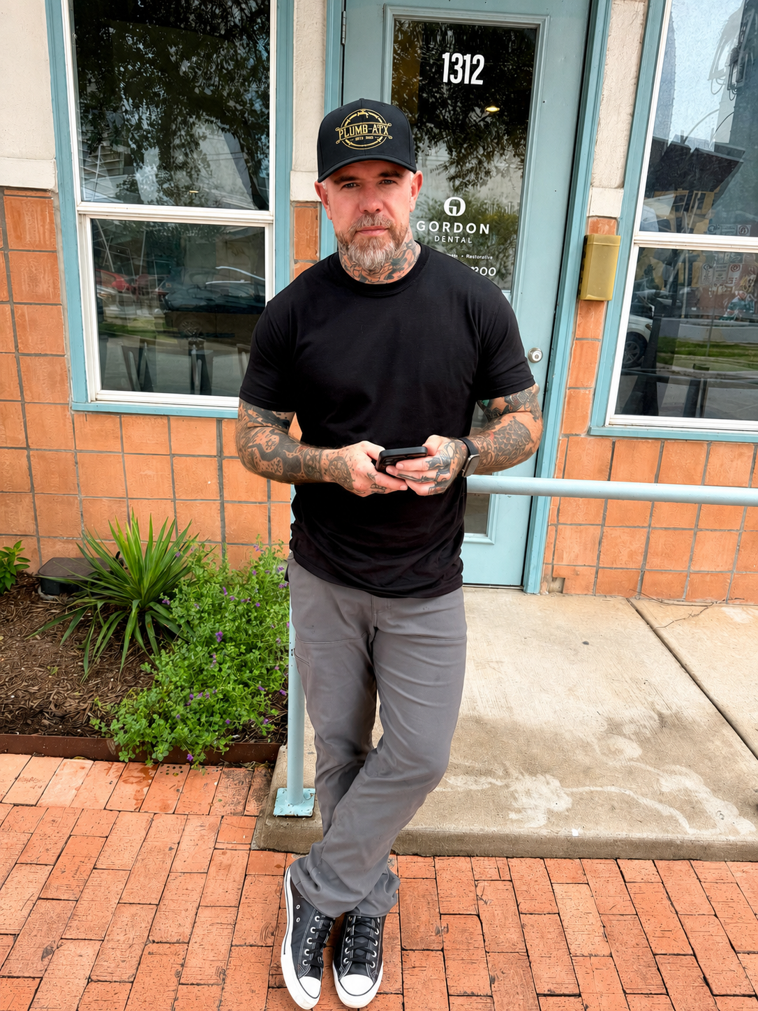 A man with tattoos wearing a black baseball cap, black t-shirt, gray pants, and black sneakers stands outside a building with a turquoise door and brick walls, holding a smartphone.