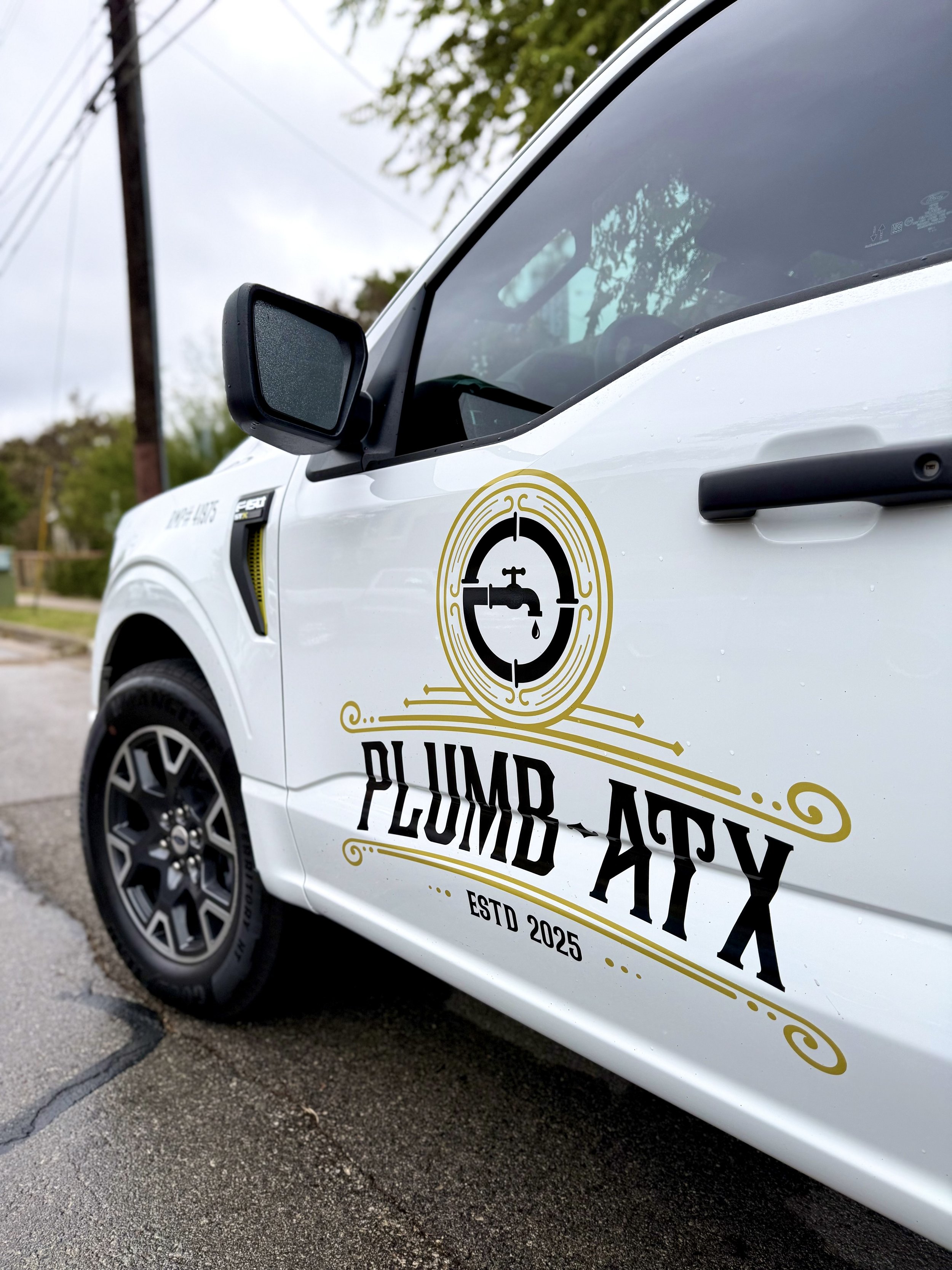 A white vehicle with a Plumbing TX logo on the door, parked on a wet street with trees and power lines in the background.