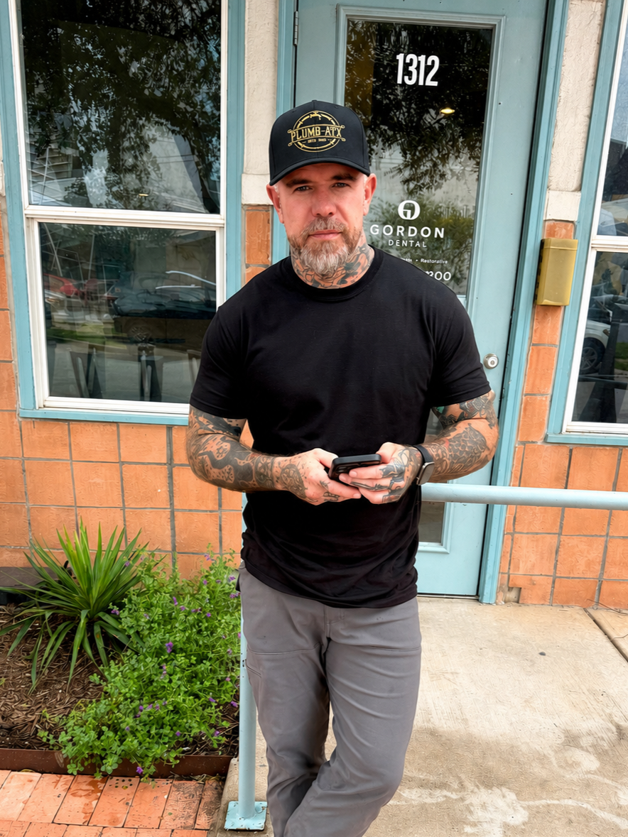 Man with tattoos wearing a black cap and black t-shirt standing outside a building, looking at the camera, holding a smartphone.