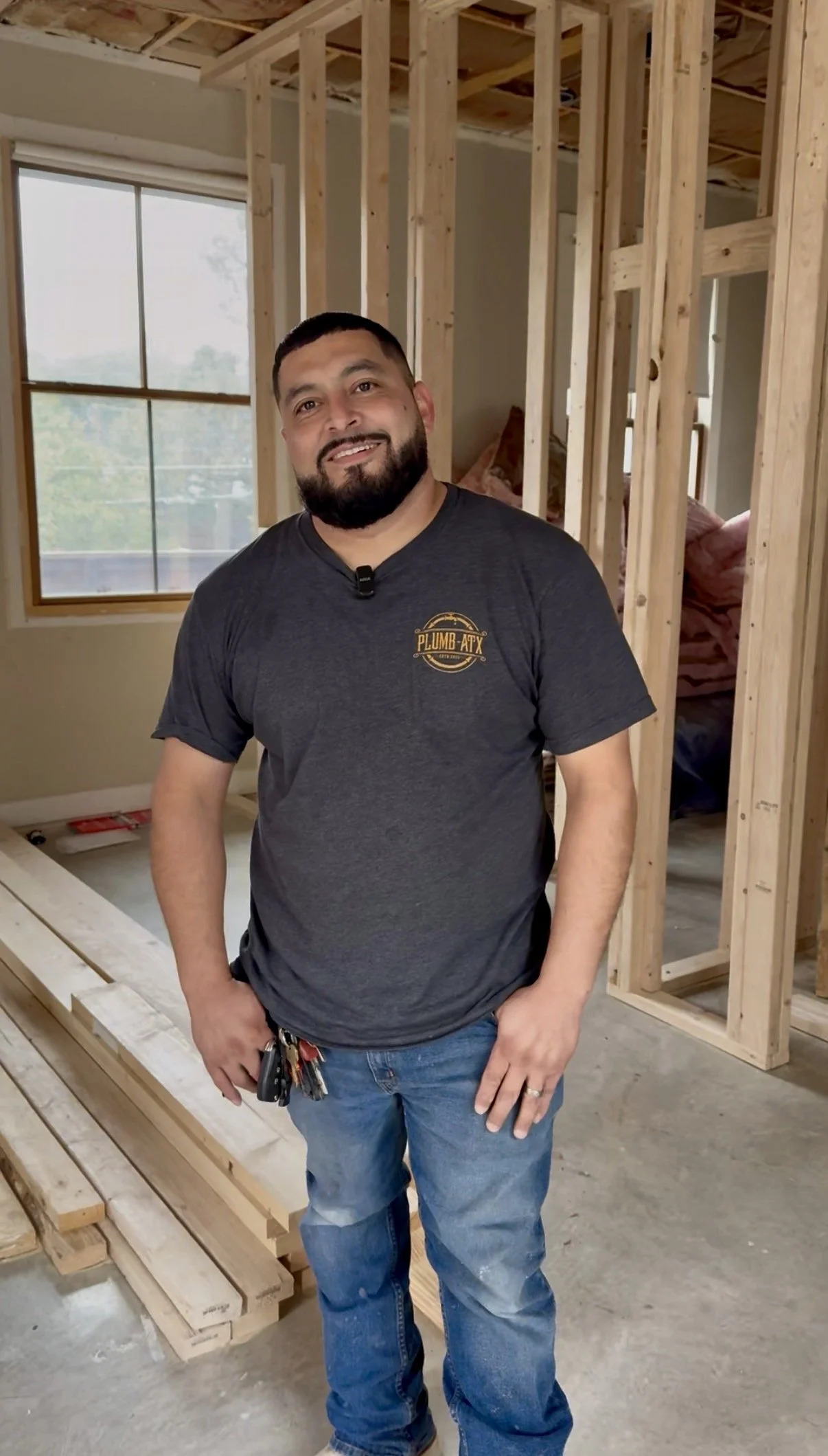 Man standing inside a house under construction, with exposed wooden framing around him and a large window in the background.