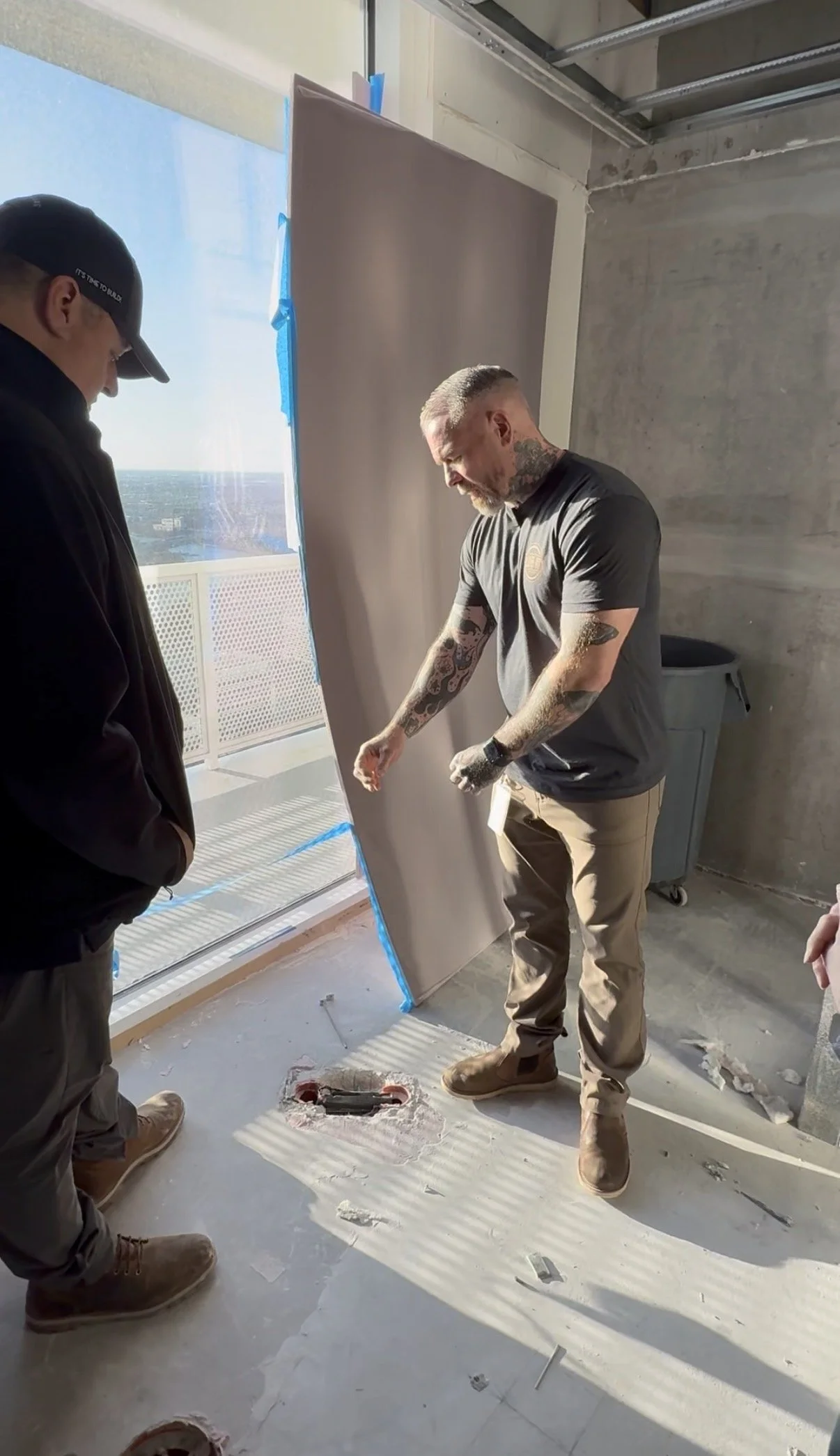 Two men are working on a construction project indoors. One man, with tattoos on his arms, is holding a large sheet of material, while the other man observes. There's a floor hole surrounded by drywall debris, and sunlight is coming through a nearby window.